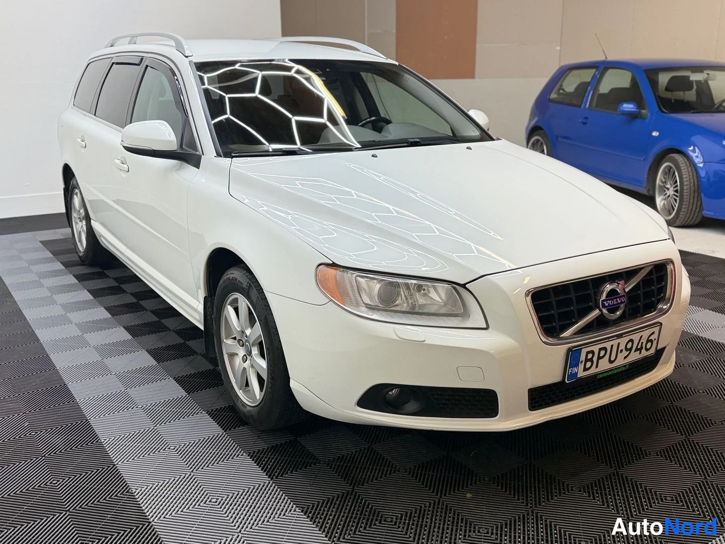 Etuviisto, nokka oikealle: Valkoinen Volvo V70 farmari kolmivartin etunurkkakuvassa sisätilassa, rekisterikilpi, kattokaiteet ja tuuliohjaimet näkyvissä.