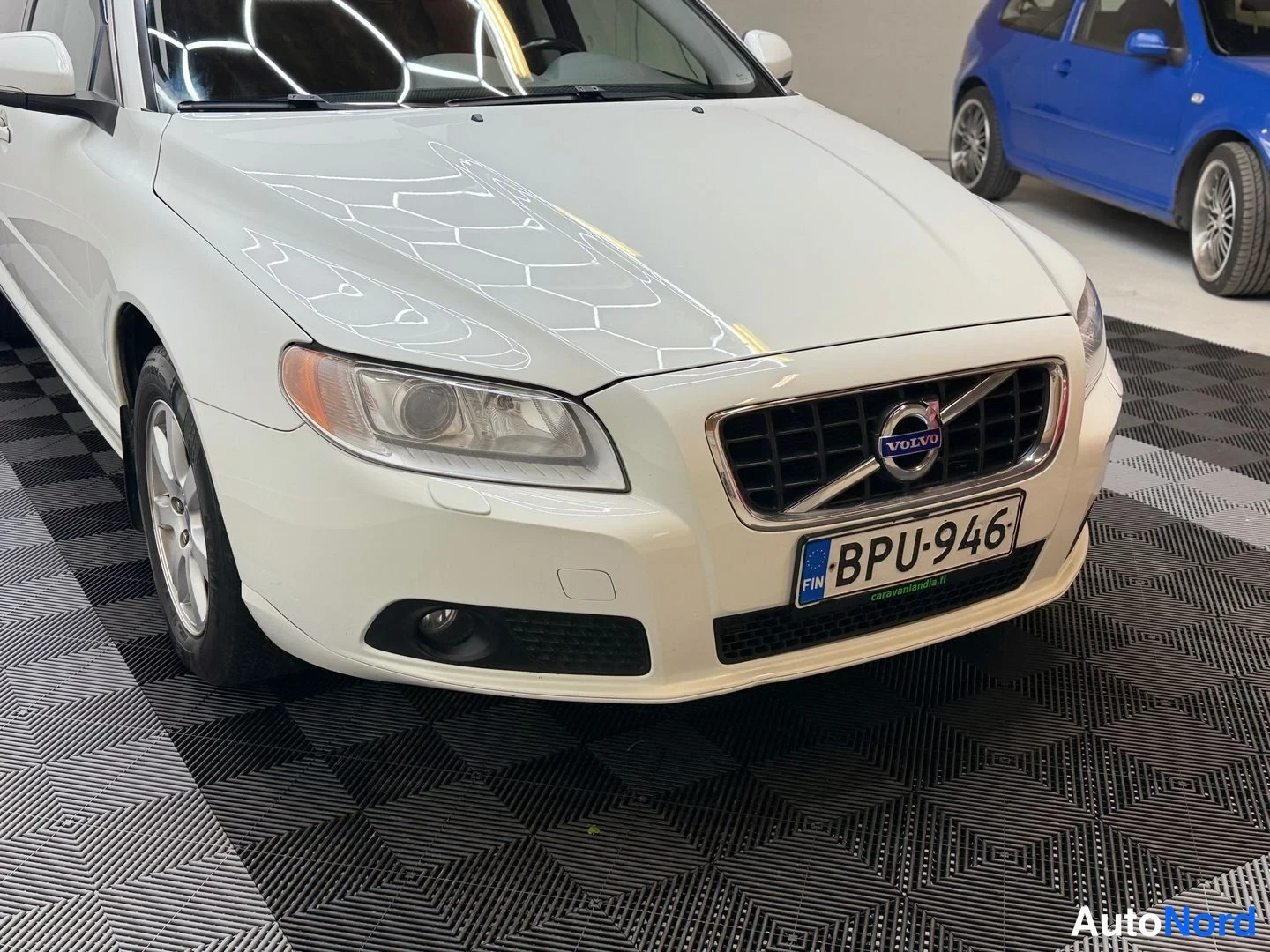 Edestä: Valkoisen auton keulan lähikuva sisätilassa: etusäleikössä Volvo-merkki ja rekisterikilpi.