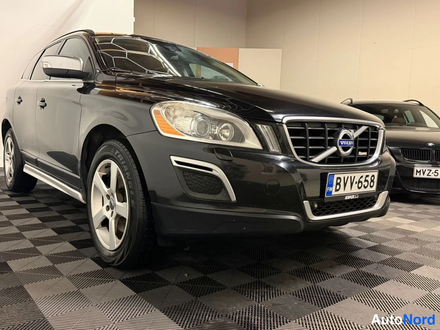 Etuviisto, nokka oikealle: Mustan tila-auton keulan ja kyljen kolmasosakulma: etusäleikkö Volvo-emblemillä, -rekisteri ja hopeiset riippumattot.