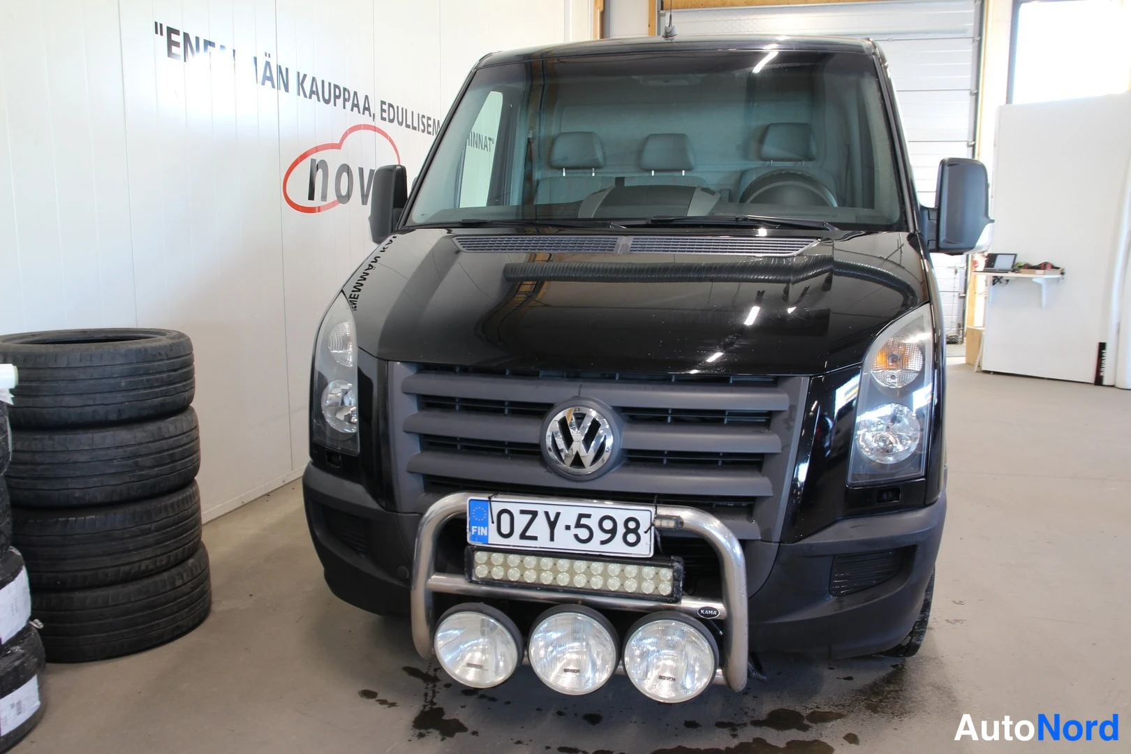 Edestä: Mustan pakettiauton etunäkymä, näkyvät VW-merkki, etusäleikkö, ajovalot, sumuvalosarvi ja -rekisterikilpi.