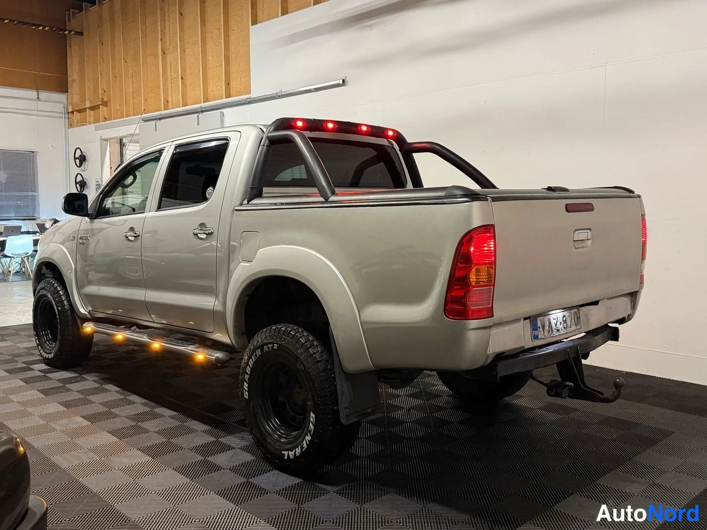 Takaviisto: Hopeanvärinen Toyota Hilux -avolava takaviistosta oikealta; lavan kansi, takavalot, rekisterikilpi ja vetokoukku näkyvät.