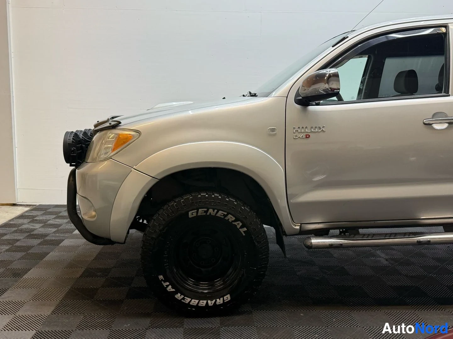 Etuviisto, nokka vasemmalle: Hopeanharmaan Toyota Hilux -avolavan etukulma: headlight, bull bar and raised suspension with General Grabber AT3 tyre visible