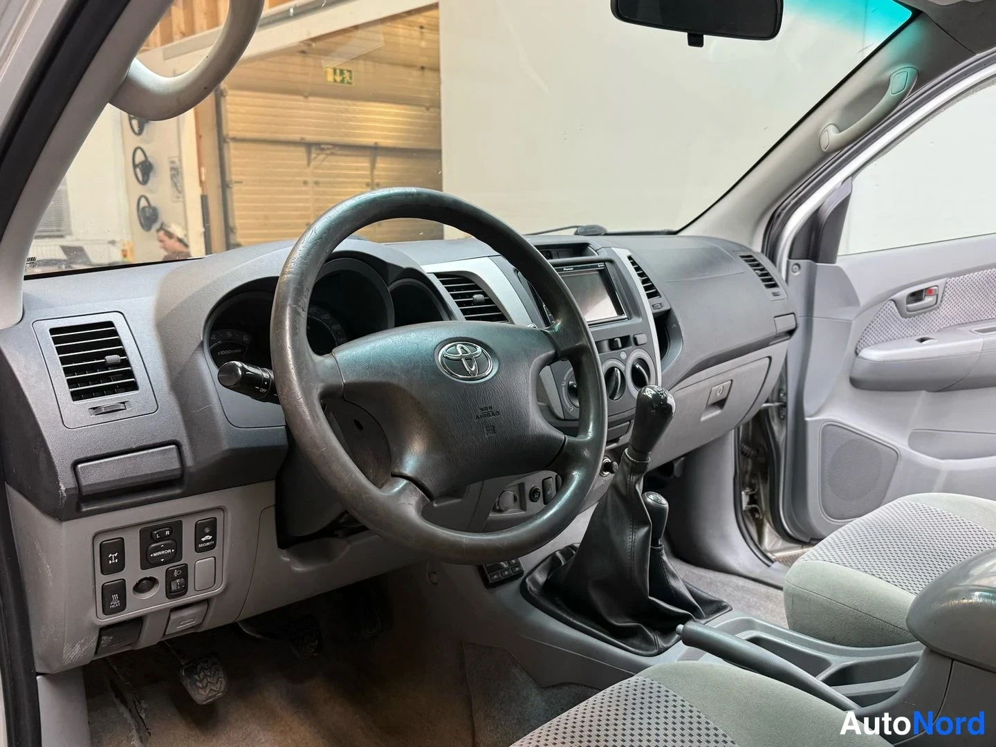 Edestä: Ohjaamon näkymä: Toyota-merkkinen ohjauspyörä, käsivaihteisto ja kojelauta.