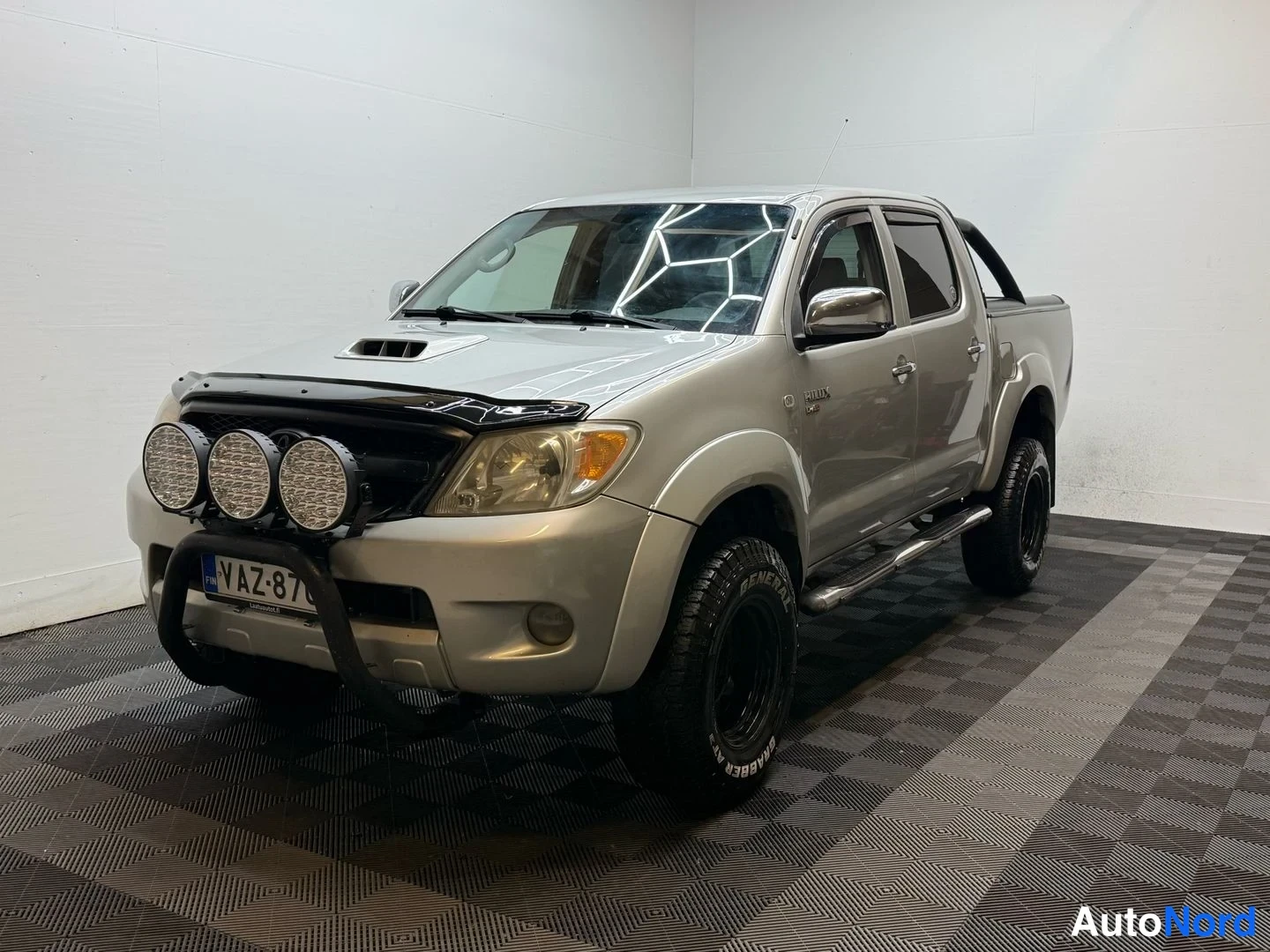 Etuviisto, nokka vasemmalle: Hopeanvärinen Toyota Hilux -avolava etuviistosta vasemmalta; keulakaari lisävaloilla, rekisterikilpi ja sivutelineet näkyvät.