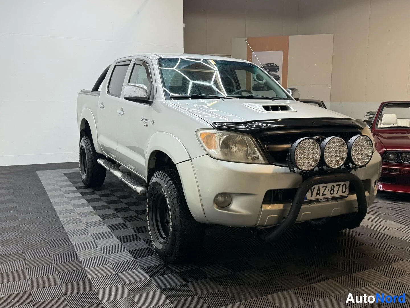 Etuviisto, nokka oikealle: Hopeanvärinen Toyota Hilux -avolava etuviistosta oikealta; keulakaari lisävaloilla, rekisterikilpi ja sivutelineet näkyvät.