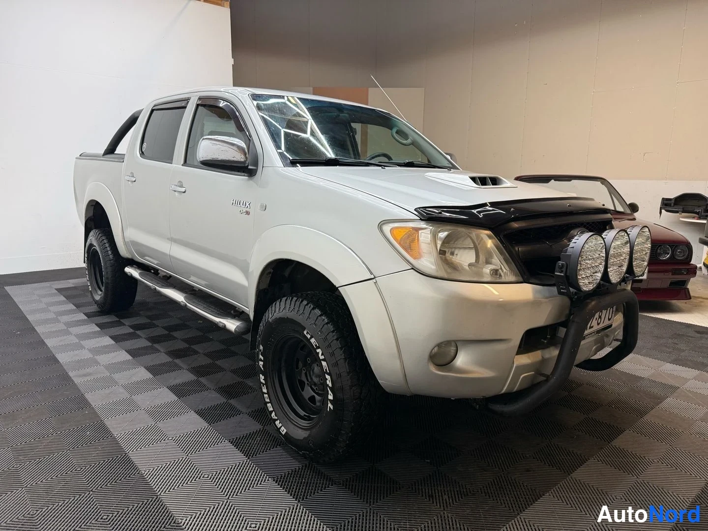 Etuviisto, nokka oikealle: Toyota Hilux -pakettiauton etukulmanosto, hopeanharmaa korimus ja mustat maastorenkaat, etupuskurissa kolmen lisävalon teline ja A-kirjain.