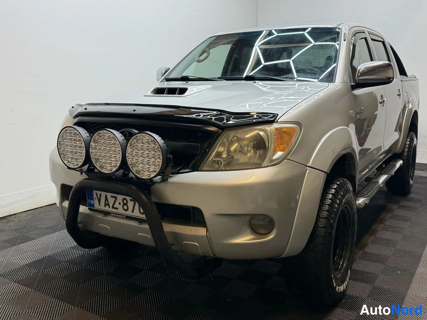 Etuviisto, nokka vasemmalle: Toyota Hiluxin etukulmanosto vasemmalta, konepellillä tuuliovi ja etupuskurin suoja; rekisterikilpi; konepellissä ilmastointijärjestelmän kyltti.