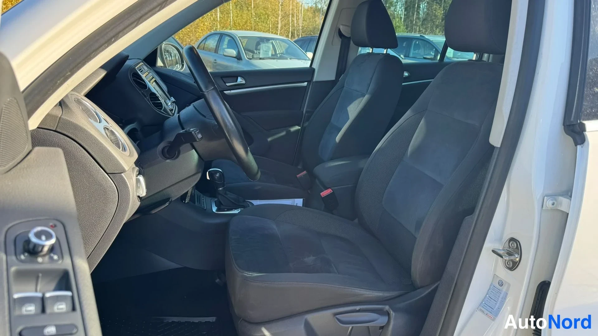 Muu: Auton sisätilat kuljettajan puolen ovesta kuvattuna: tummat kankaankupit, ohjauspyörä ja keskärmisto, valkoinen korin reuna.