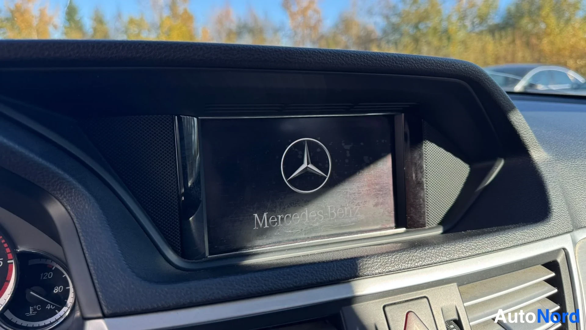 Yksityiskohta: Kojelaudan upotetun näytön käynnistysruutu, jossa Mercedes-Benzin tunnus ja teksti Mercedes-Benz. Vasemmalla näkyy osa ratti- ja mittaristosta.