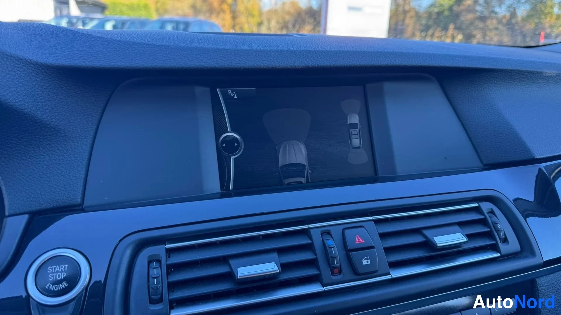 Muu: Auton kojelaudan näyttö, jossa näkyy peruutuskameran tai pysäköintitunnistuksen kuva ja ajoneuvon grafiikka; alla ilmastointiaukot ja Start/Stop Engine -painike