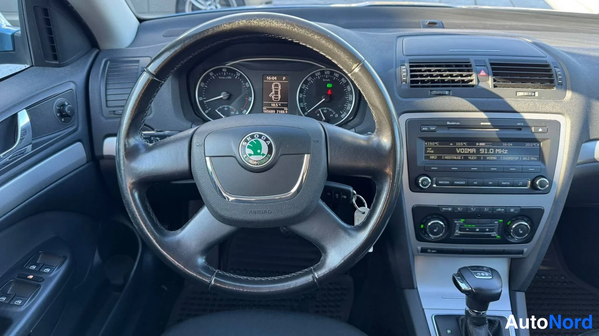 Edestä: Ohjauspyörä lähikuvassa Skoda-logolla, takana kojelaudan mittarit, radio ja automaattivaihteiston valitsin; vasemmalla oven sähköikkunakytkimet.