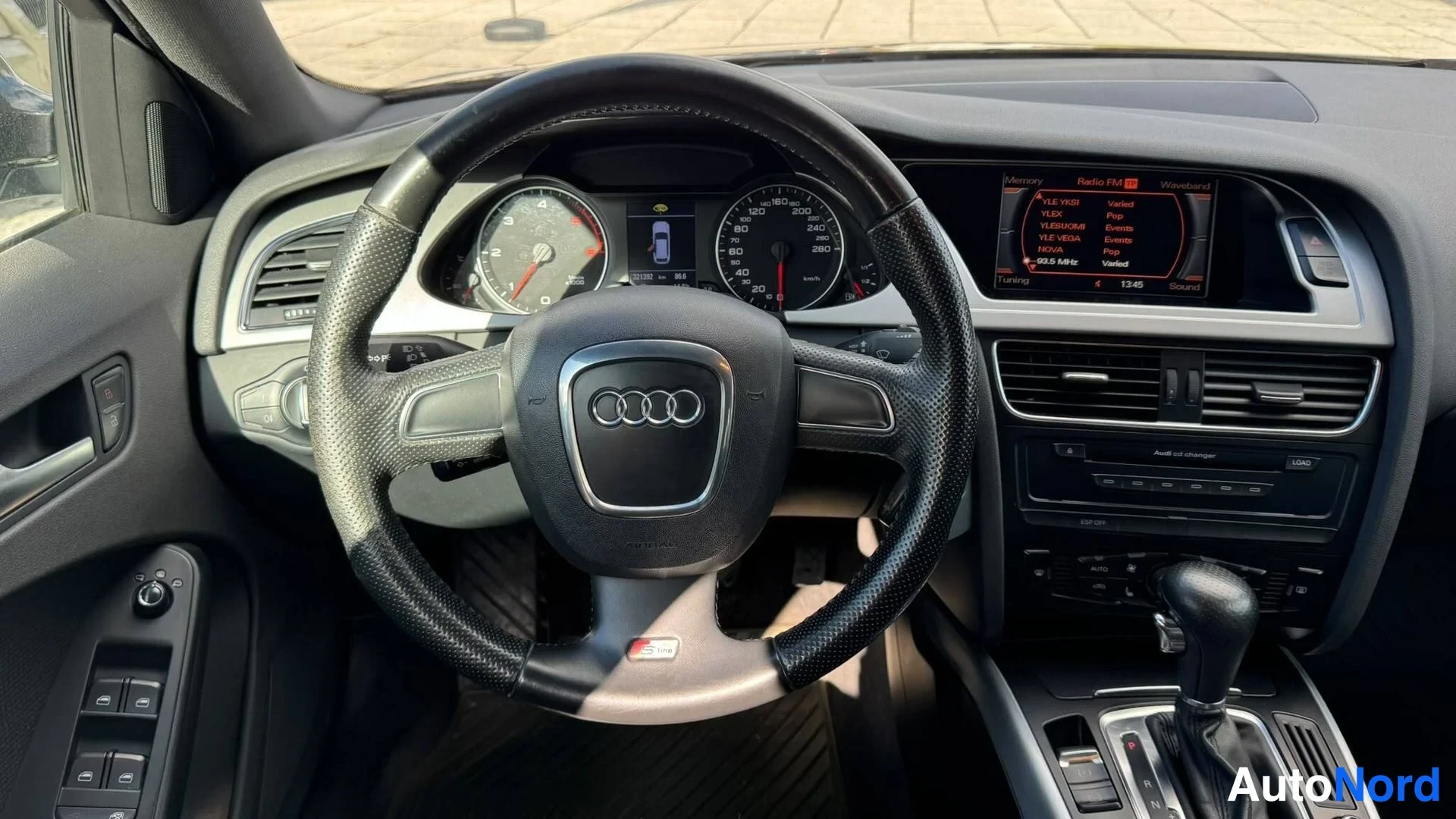 Muu: Ohjauspyörä lähikuvassa, Audi-tunnus ja S line -merkki pyörän nápissa, mittaristo ja keskikäyttötaulu näkyvissä.