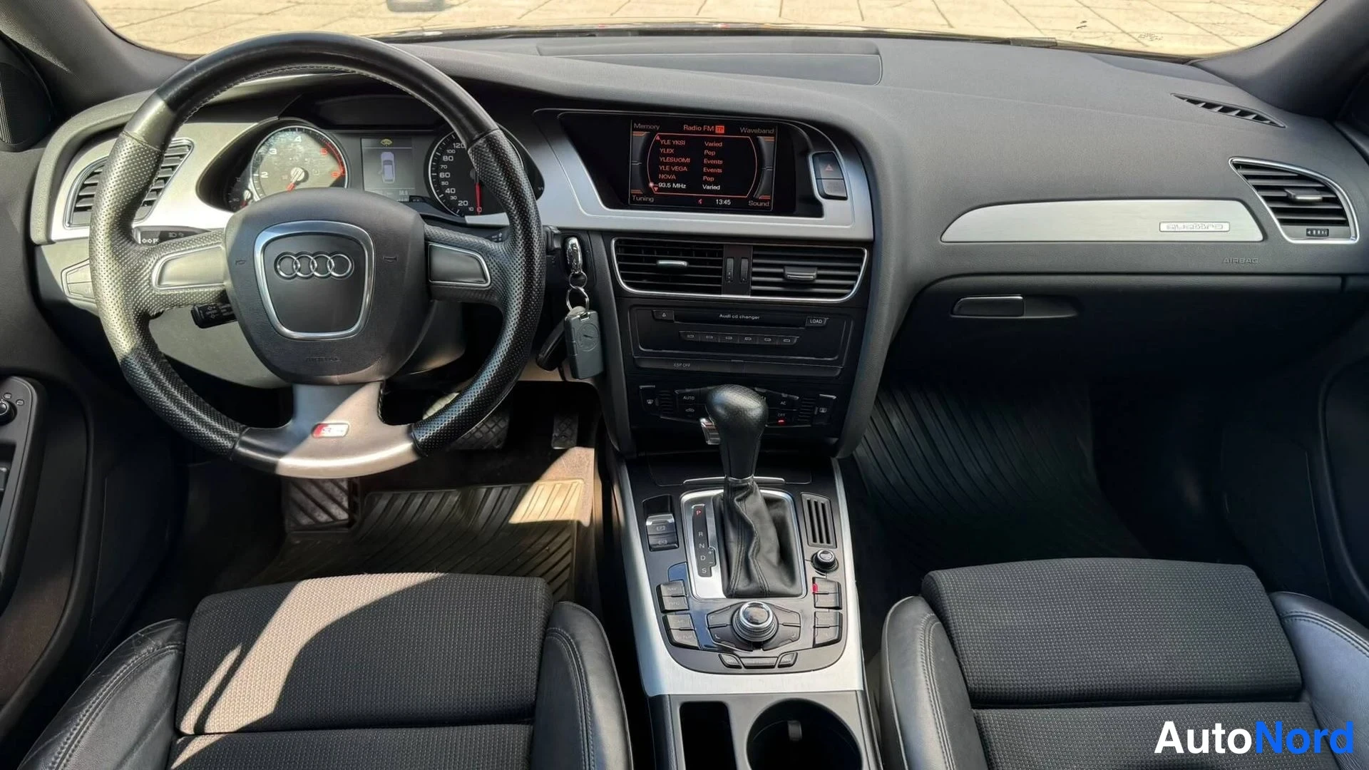 Muu: Ohjaamo etuosasta nähtynä: Audi-merkkinen ohjauspyörä, automaattivaihteen valitsin, kojelaudan näyttö ja radio, mustat istuimet.