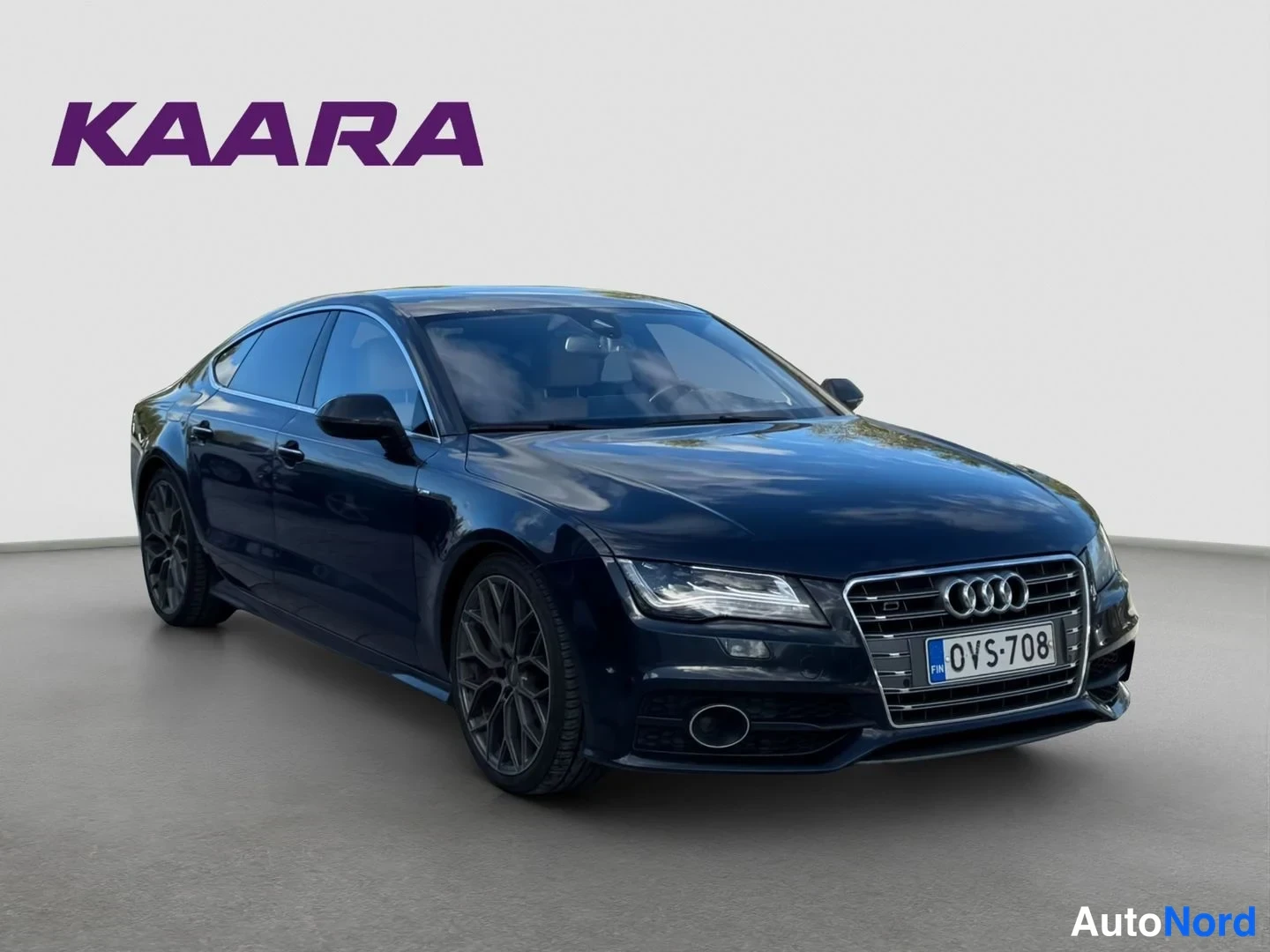Etuviisto, nokka oikealle: Audi A7 -auto tummansinisenä etukulmasta oikealle, rekisteritunnus, studiossa harmaata lattiaa vasten.