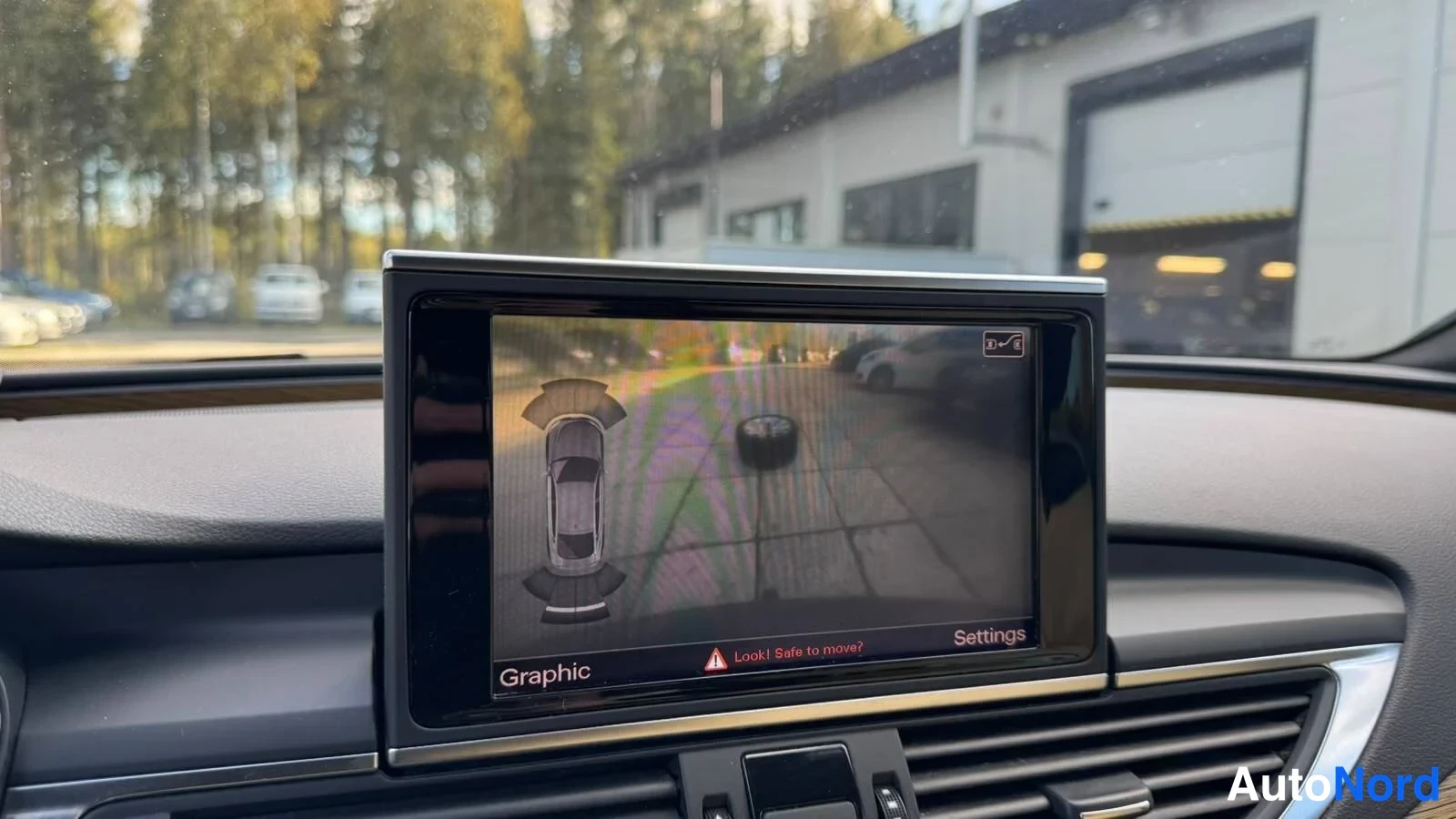 Muu: Audi A7:n keskikonsolin näyttö, jossa näkyy peruutuskameran kuva ympyräviivausmerkinnöineen ja tekstit Graphic, Settings ja Lock! Safe to move?