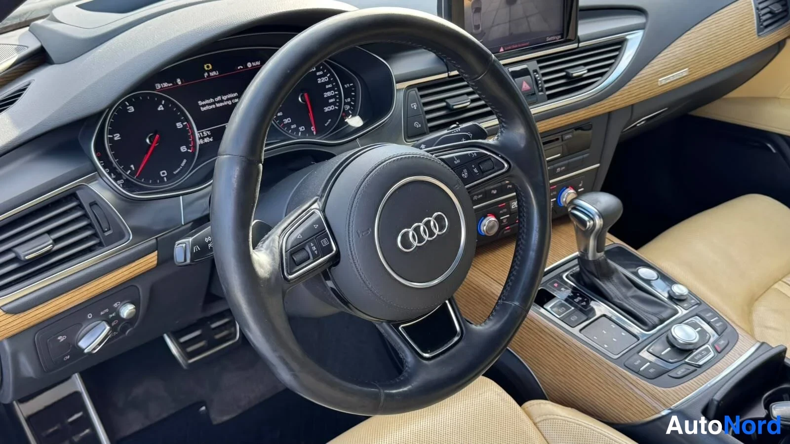 Etuviisto: Audi A7:n ohjaamo: Audi-merkkinen ohjauspyörä, mittaristo, MMI-näyttö ja parkkihälytyspainikkeet keskitunnistossa, vaaleanruskea nahkapinta ja puunäköinen koris