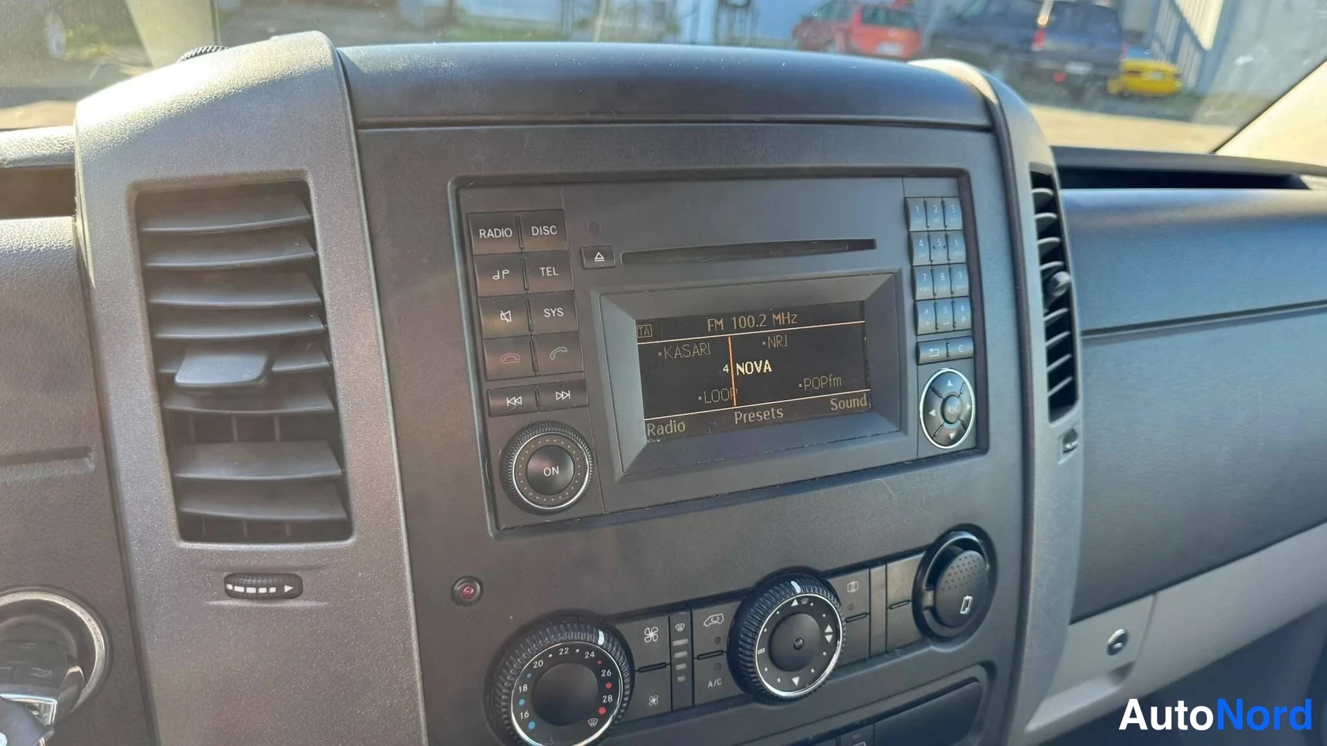 Muu: Kojelaudan keskiosa lähikuvassa: radio- ja CD-yksikkö, jonka näytöllä on käynnissä FM 100.2 MHz ja 'NOVA', sekä alla olevia säätöpainikkeita.