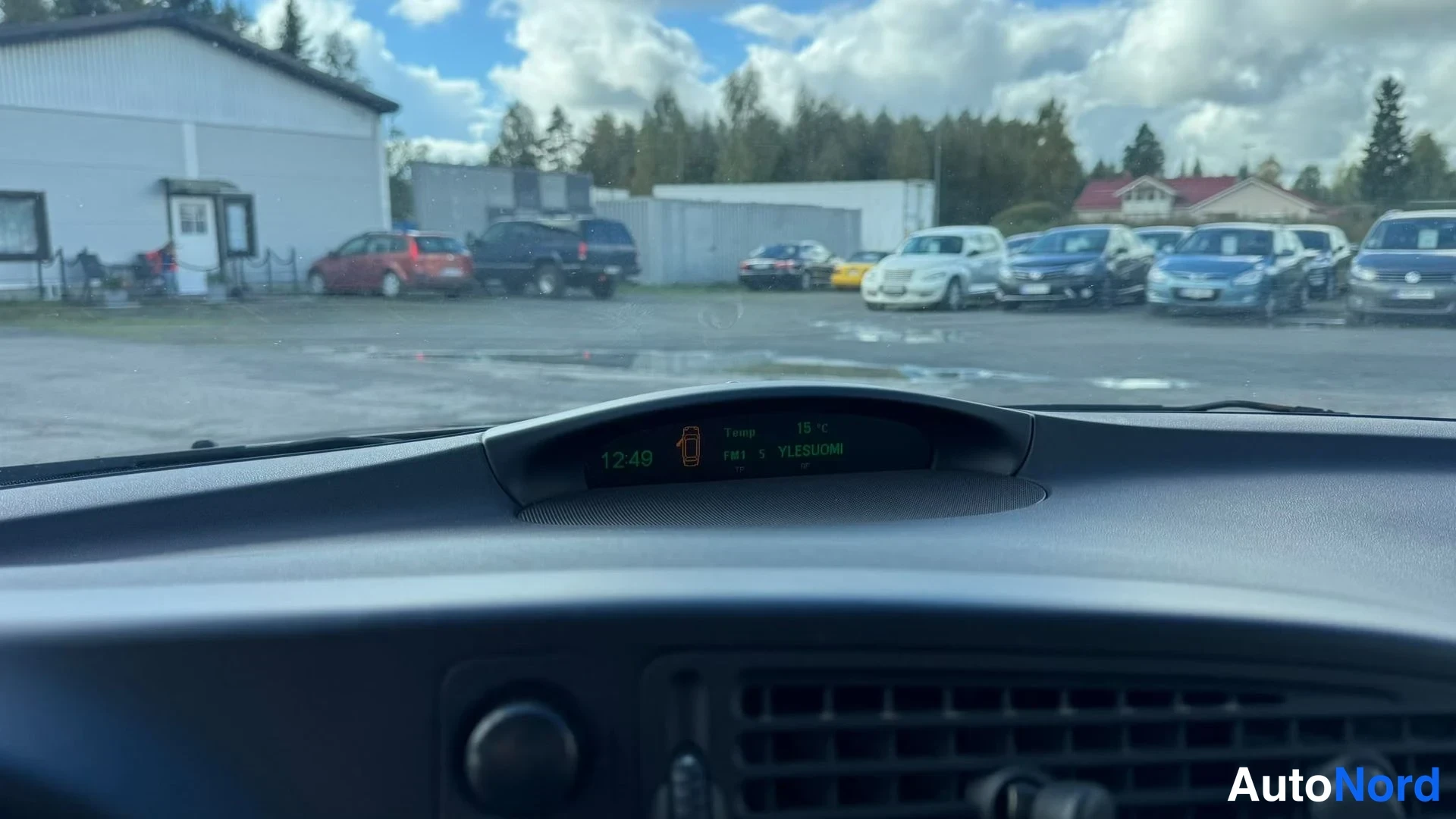 Muu: Saab 9-3:n kojelaudan yläosa ja tuulilasista näkyvä parkkipaikka; infonäytössä kello 12:49, lämpötila 19 °C ja FM1 5 YLESUONI.