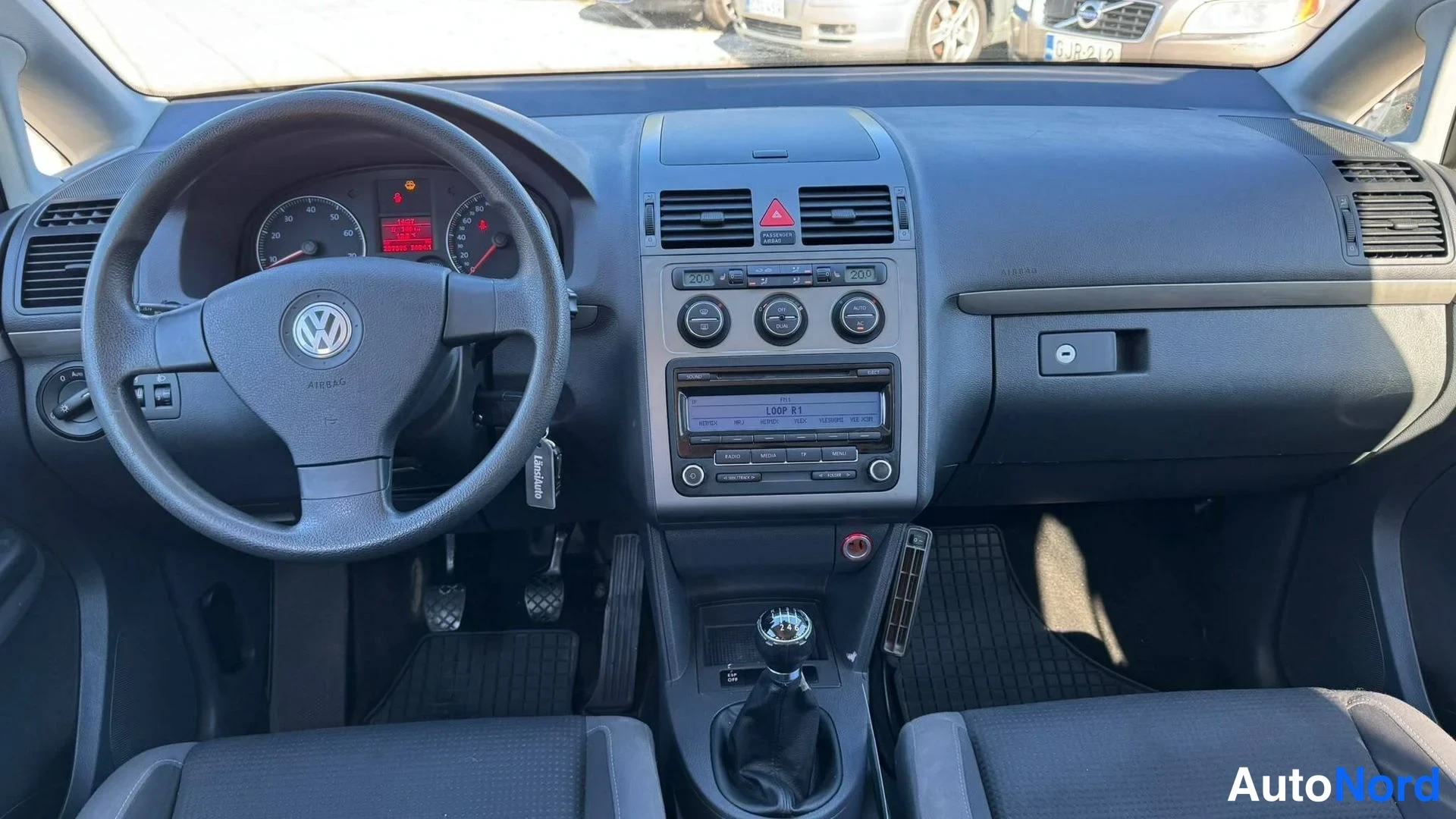 Muu: Ohjaamo edestä kuvattuna: VW-ohjauspyörä, manuaalivaihtopylkäs, radio ja digitaalinen näyttö kojelaudassa.