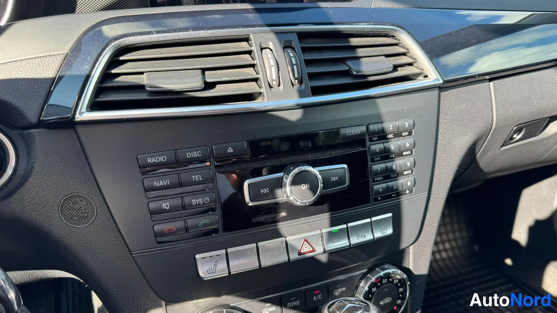 Muu: Keskeiskonsolin radio- ja nappipaneeli lähikuvassa: RADIO, DISC, NAVI, TEL- ja SYS-napit, numeronäppäimistö ja sulautetut ilmastointilaitteet.