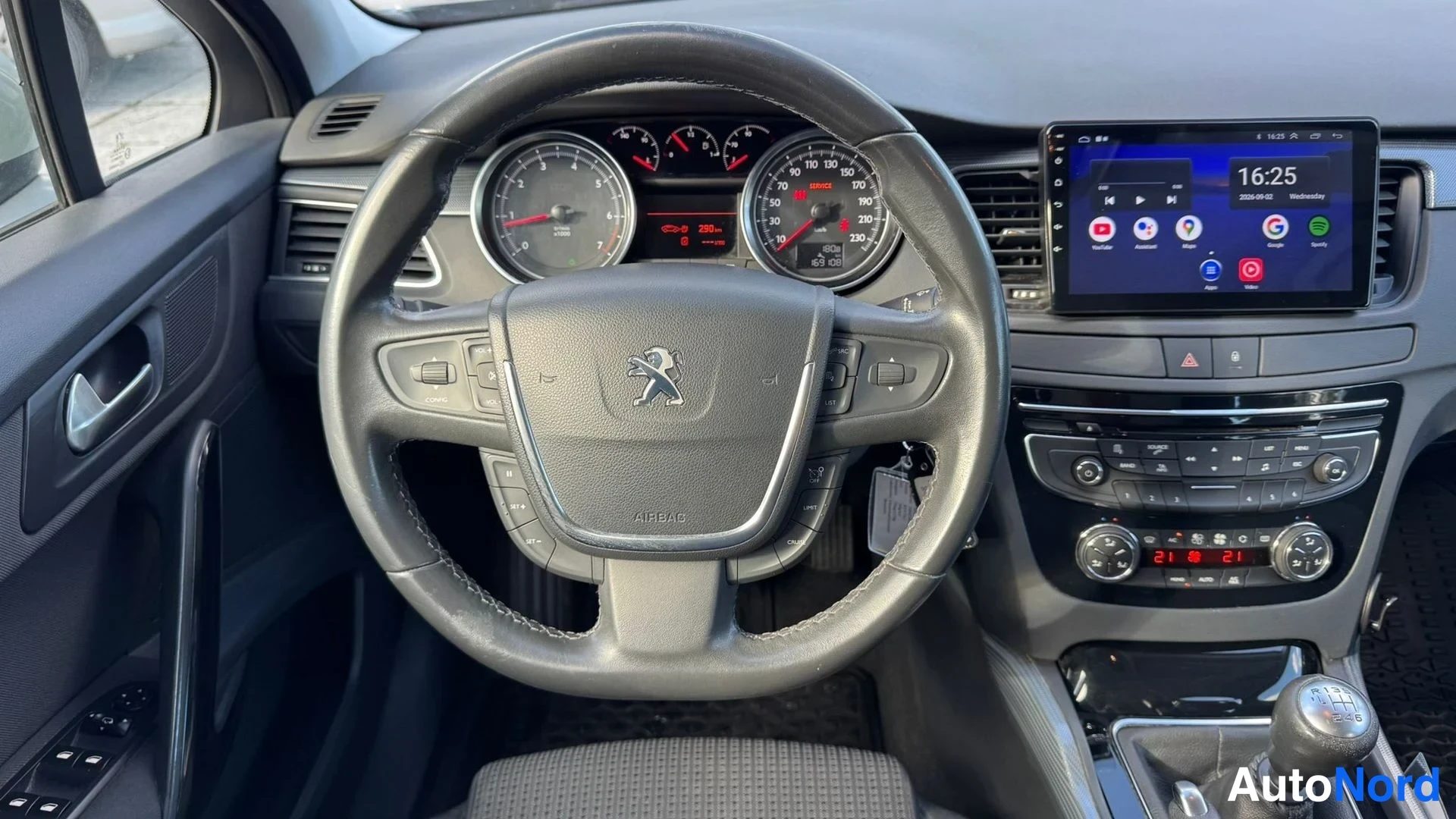 Edestä: Ohjauspyörän lähikuva Peugeot-logolla ja AIRPACK-tunnisteella, taustalla konepelti, moottorin lämpömittari, multimediaosasto ja tumma kojelauta.