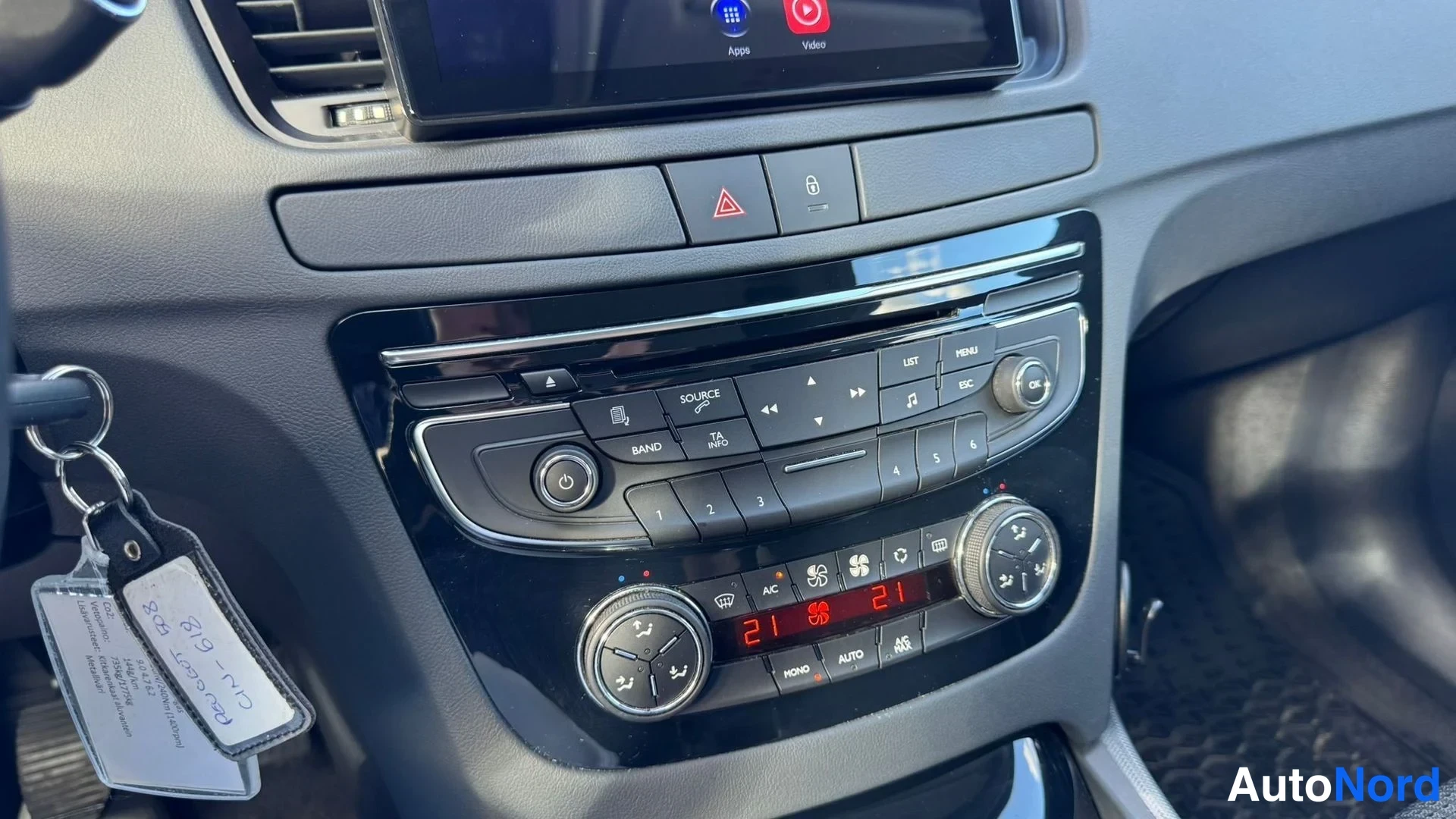 Yksityiskohta: Keskitunnin radio- ja ilmastointipaneeli lähikuvassa; ilmastointi asetettu 21 asti, näkyvissä AUTO- ja A/C-napit sekä virta- ja lähdekuvakkeet. Vasemmalla avaim