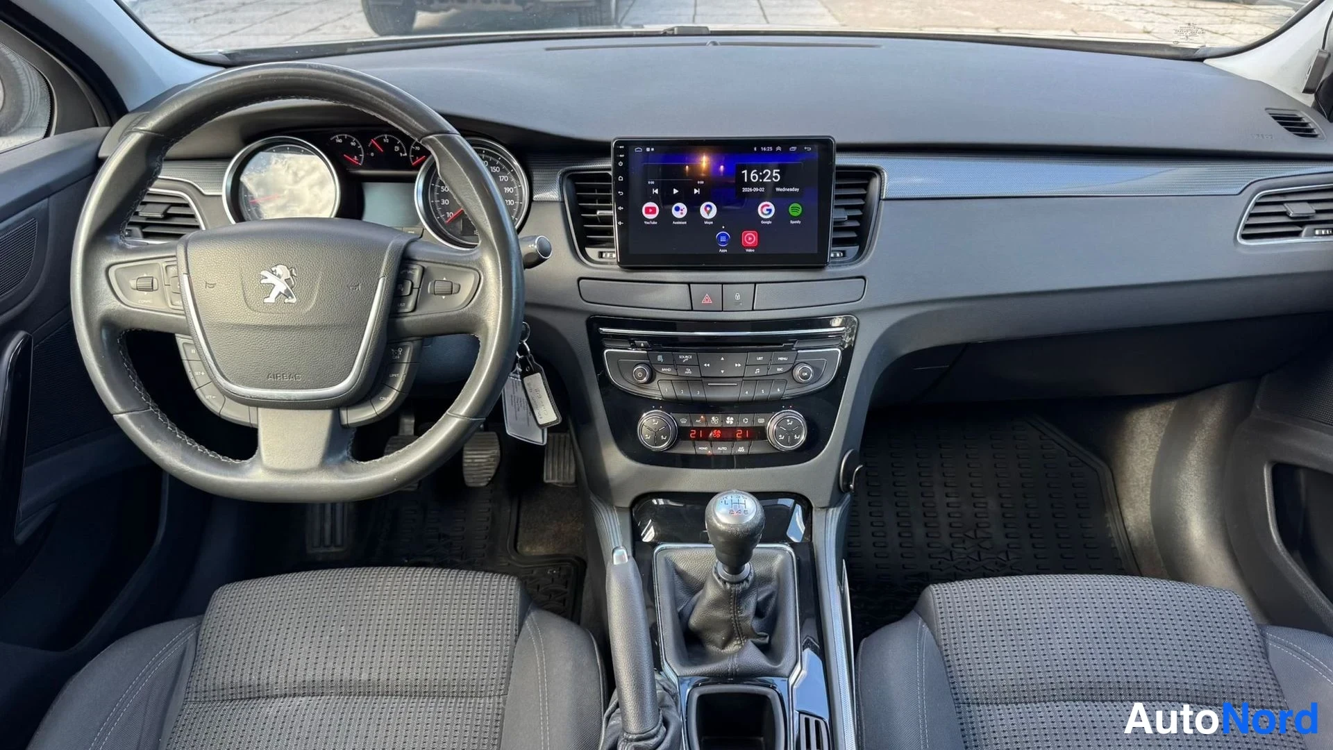 Edestä: Ohjaamo etupuolelta: tumma kojelauta, monitoimiohjaus pyörässä Peugeot-logolla, keskitunnin suuri kosketusnäyttö, manuaalinen vaihtelaakeri ja kangasistuimet.