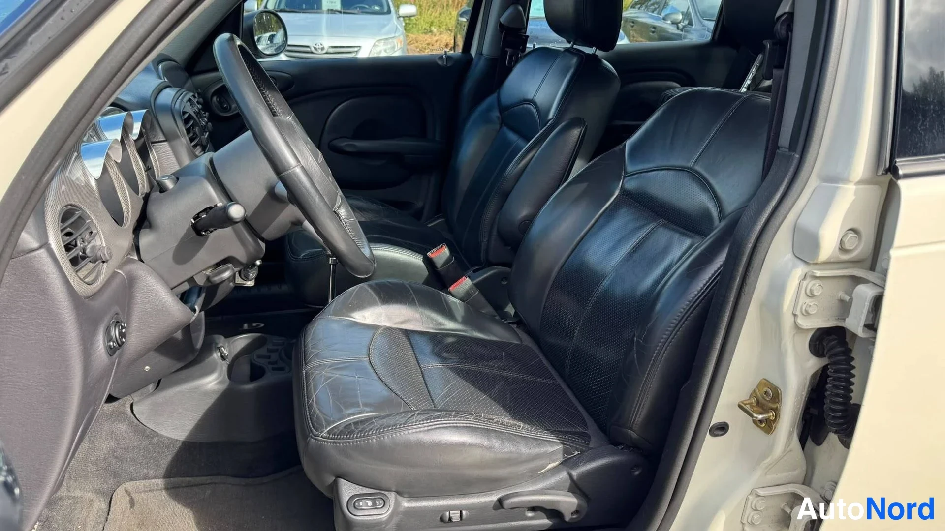 Takaa: Chrysler PT Cruiserin etutupaikkasta: musta nahkainen etuistuin, käsivarsinen nappi ohjauspyörän takana ja vasemman oven avausosa näkyvät.