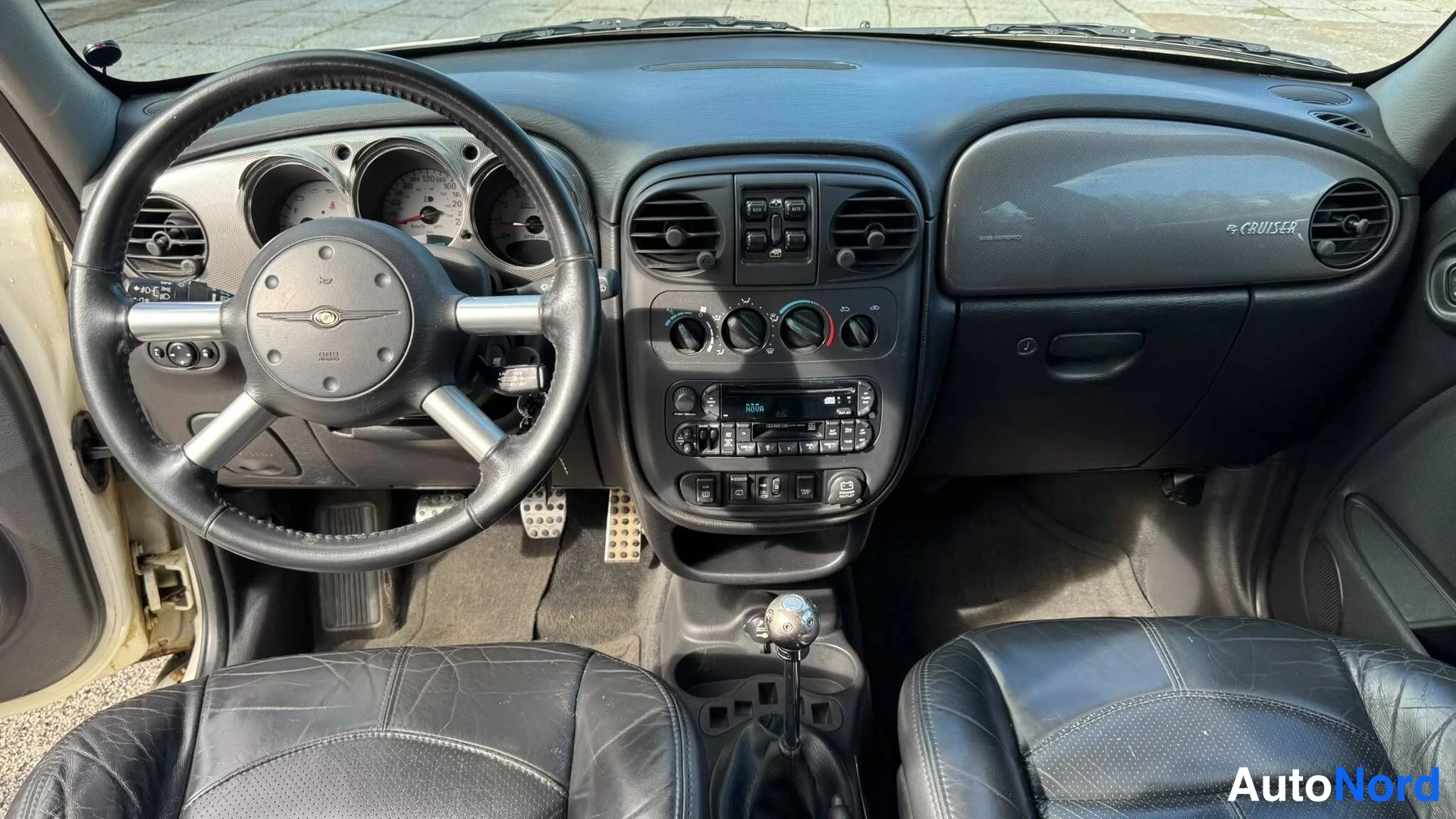 Etuviisto, nokka vasemmalle: Ohjaamo edestä: musta nahkapenkit, manuaali vaihtaja, Chrysler-merkitty ohjauspyörä ja kojelauta, jossa teksti PT CRUISER.