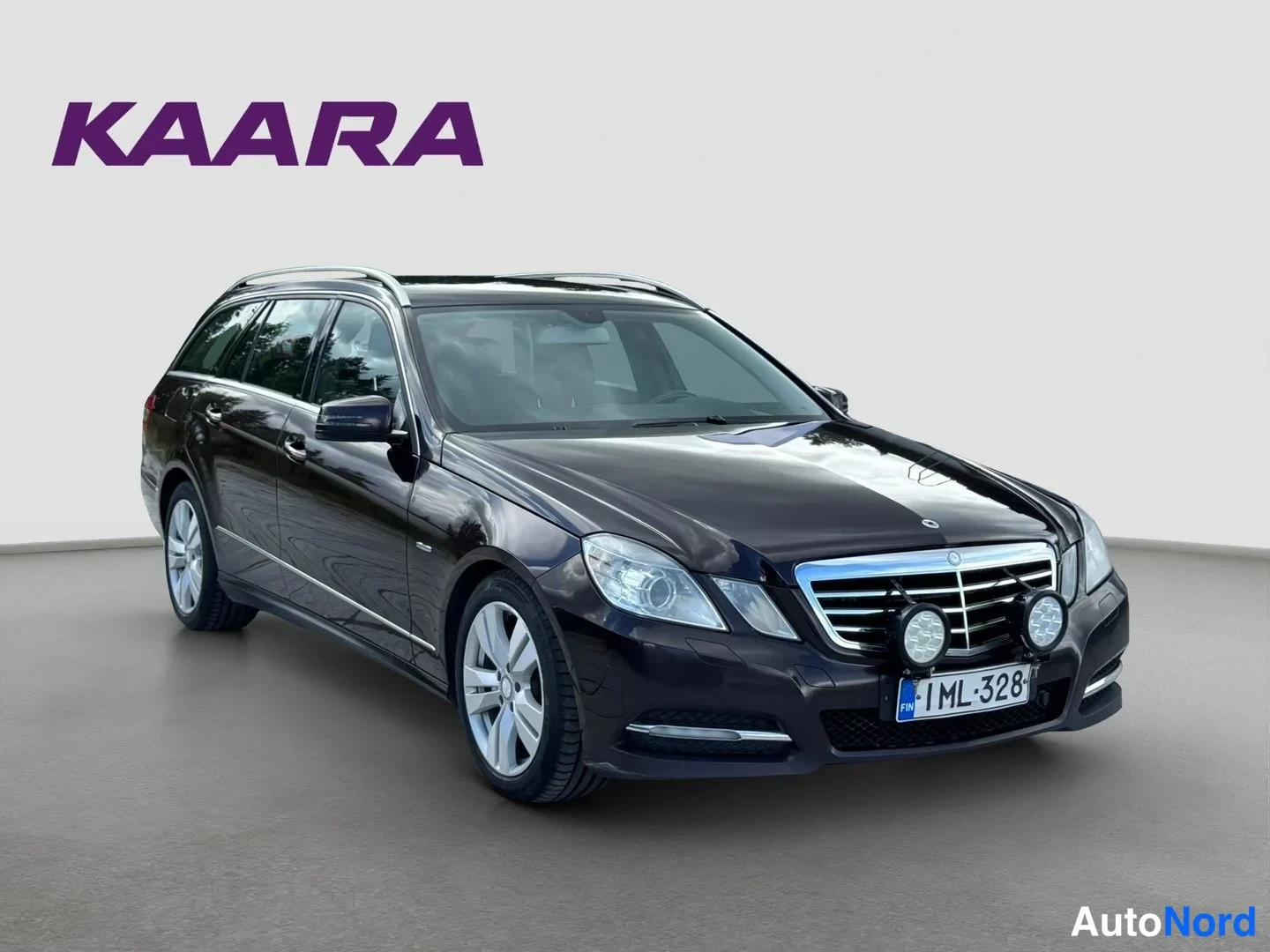 Etuviisto, nokka oikealle: Mercedes-Benz E-sarjan farmari etuviistosta oikealta, tummanruskea koriväri, etusäleikkö ja vasen kylki näkyvissä, lisävalot etupuskurissa.