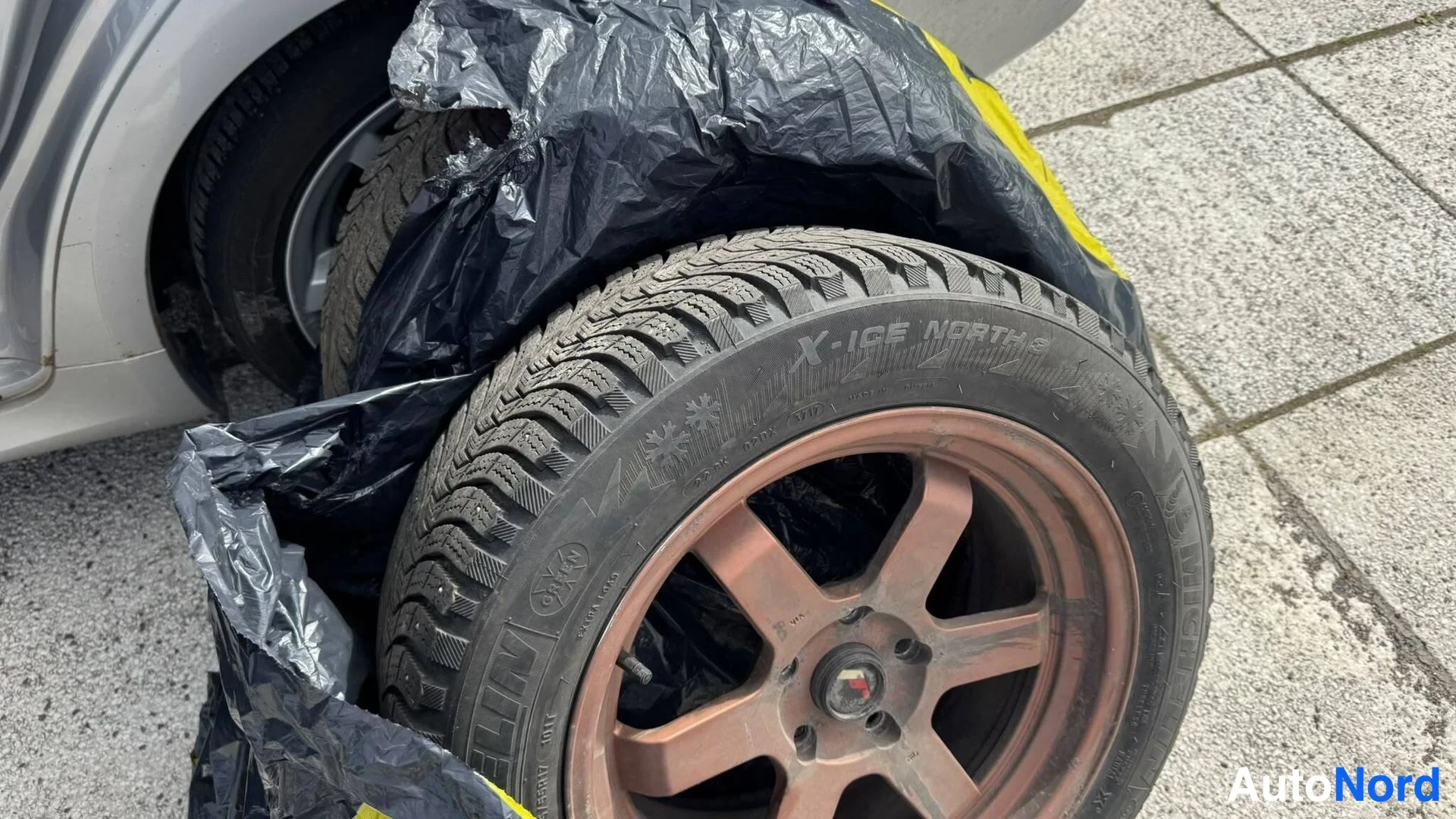 Muu: Irrotettu rengas vanteella mustan muovipussin päällä, Michelin X-Ice North talvirengas.