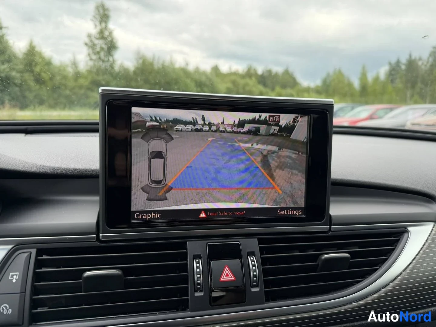 Muu: Näyttö näyttää peruutuskameran kuvan parkkipaikasta, Graphic- ja Settings-teksteillä sekä Look! Safe to move? -varoituksella.