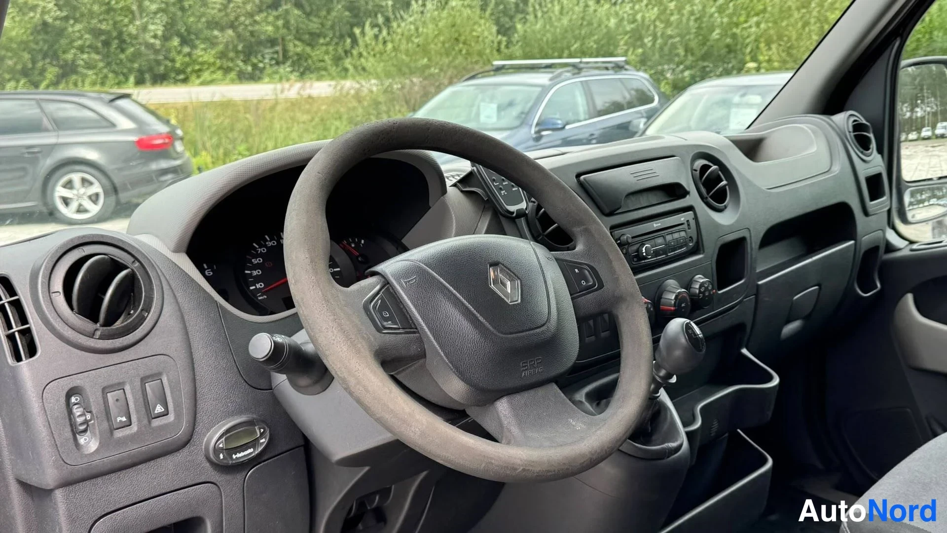 Takaviisto: Ohjaamon näkymä: tumma ohjauspyörä Renault-logolla, kojelauta ja manuaali vaihdetta.