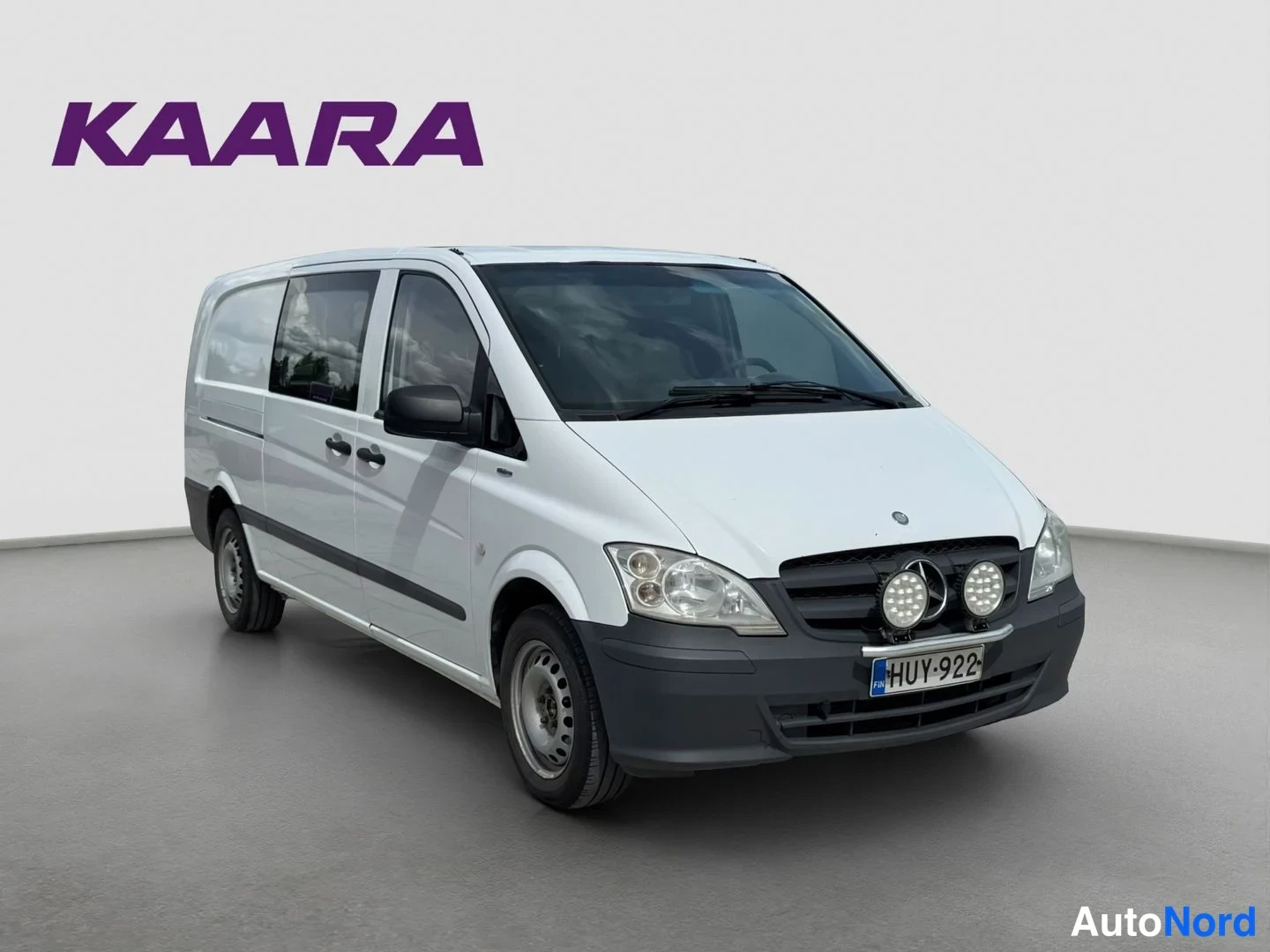 Etuviisto, nokka oikealle: Valkoinen Mercedes-Benz Vito -pakettiauto kolmesta neljäsosasta, etupää oikealla, rekisterikilpi ja kaksi lisävaloa etupuskurissa.