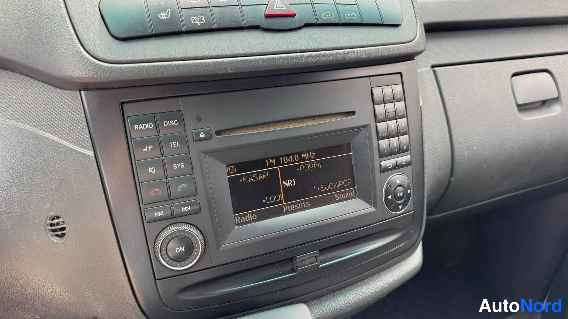 Muu: Kojelaudan keskialue: radiopainikkeet, numeronäppäimistö ja näyttö, jossa lukee FM 104.0 MHz ja NRJ.