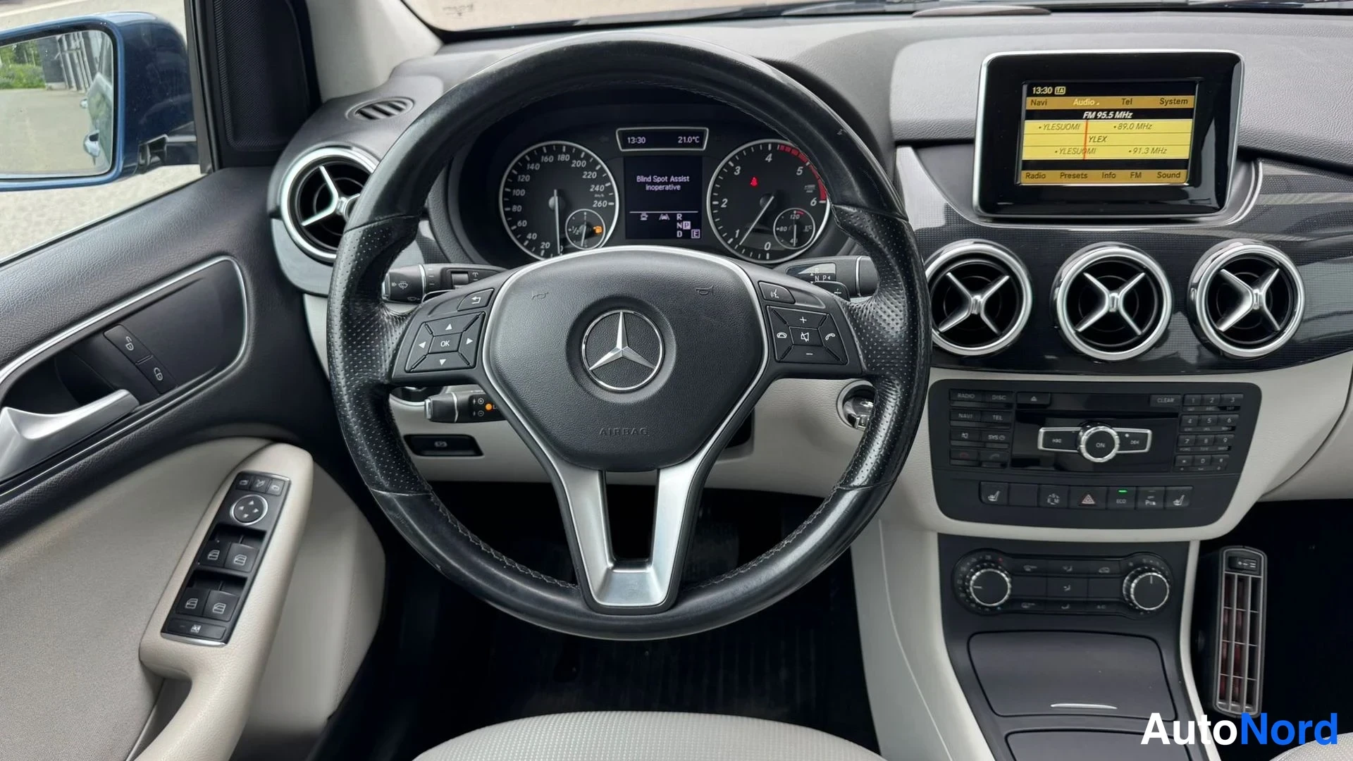 Muu: Ohjauspyörä ja mittaristo Mercedes-Benzissä, näytöllä radiovalikko, oviaukkoinstrumentit vasemmalla.
