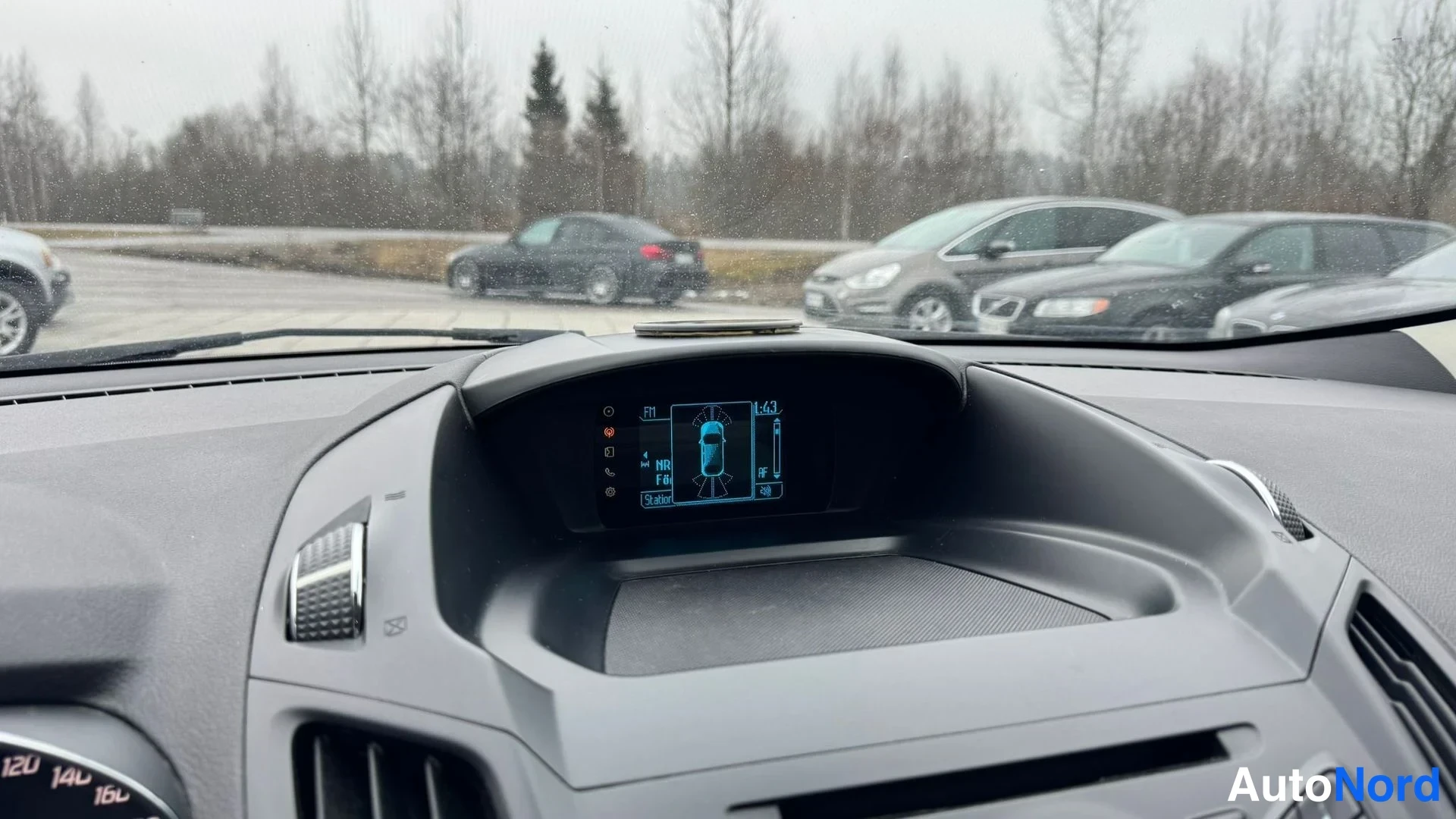 Muu: Kojelaudan yläosan infonäyttö lähikuvassa, jonka taustaa tuulilasin läpi näkyy parkkialue autoineen.