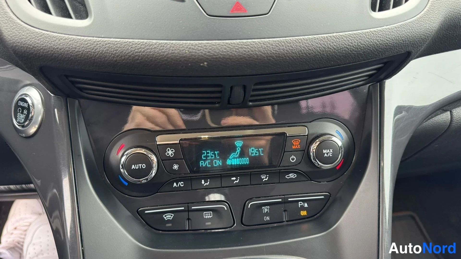Yksityiskohta: Ilmastointilaitteen hallintapaneeli, jonka näytössä lukee '23°C, 19°C, A/C ON'; näkyvissä AUTO-, A/C- ja MAX A/C -painikkeet, ENGINE START STOP -painike sekä P/