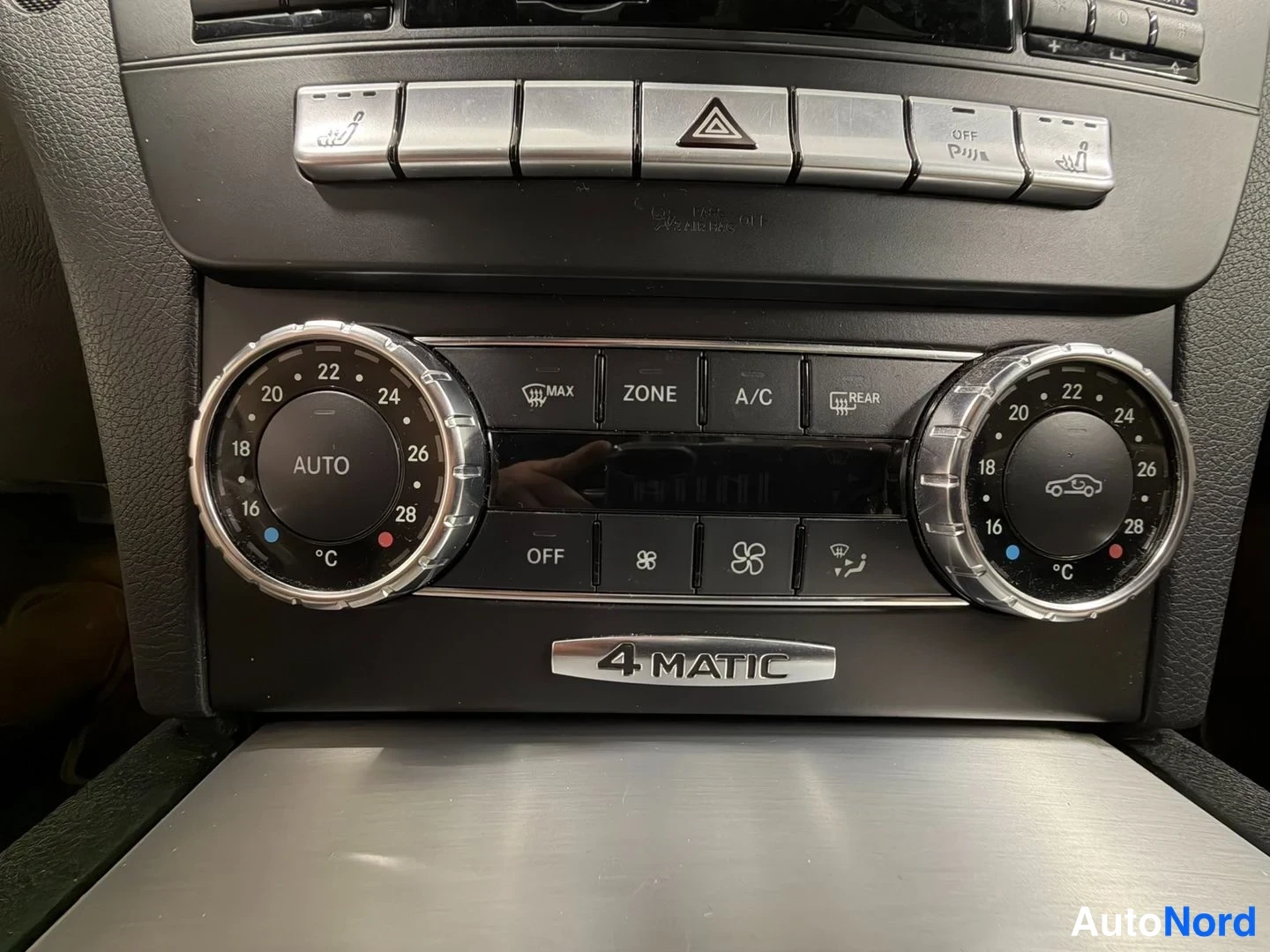 Muu: Mercedes-Benzin kojelaudan ilmastointipaneeli lähikuvassa: kaksi pyöreää säädintä AUTO-merkinnällä ja asteikolla 16–28 °C, keskellä painikkeet MAX, ZONE, A/C ja