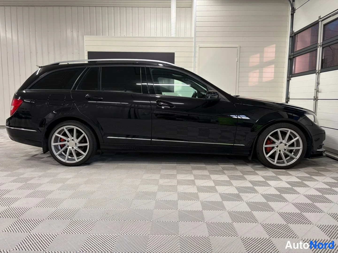 Sivulta: Mustan Mercedes-Benz farmarin sivuprofiili sisätiloissa; hopeiset kattokaiteet ja kevytmetallivanteet näkyvät.