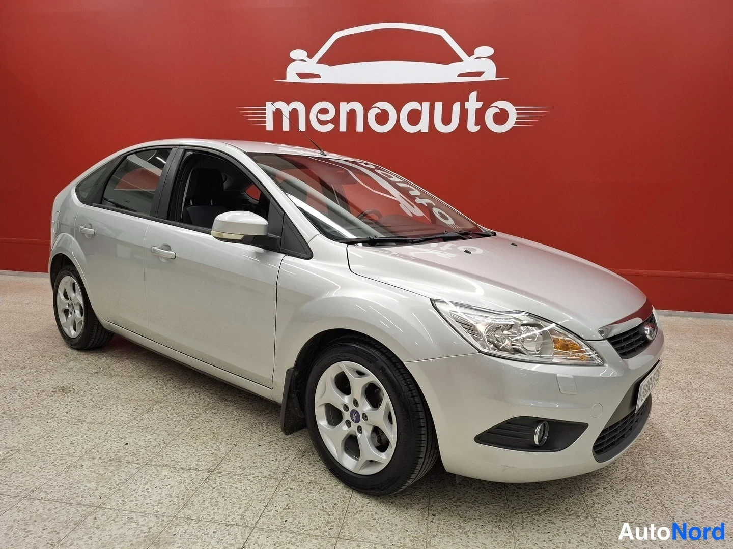 Etuviisto, nokka oikealle: Hopeanvärinen Ford Focus -viistoperä kuvattu kolmanneltyyli edestä oikealta sisätiloissa punaisella taustalla.