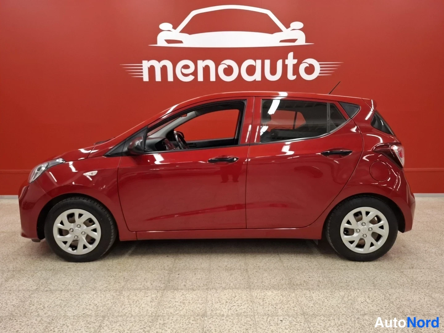 Etuviisto, nokka vasemmalle: Punainen Hyundai i10 -henkilöauto sivulta etukolmannelta kuvakulmalla sisätiloissa punaisella menoauto-taustalla.
