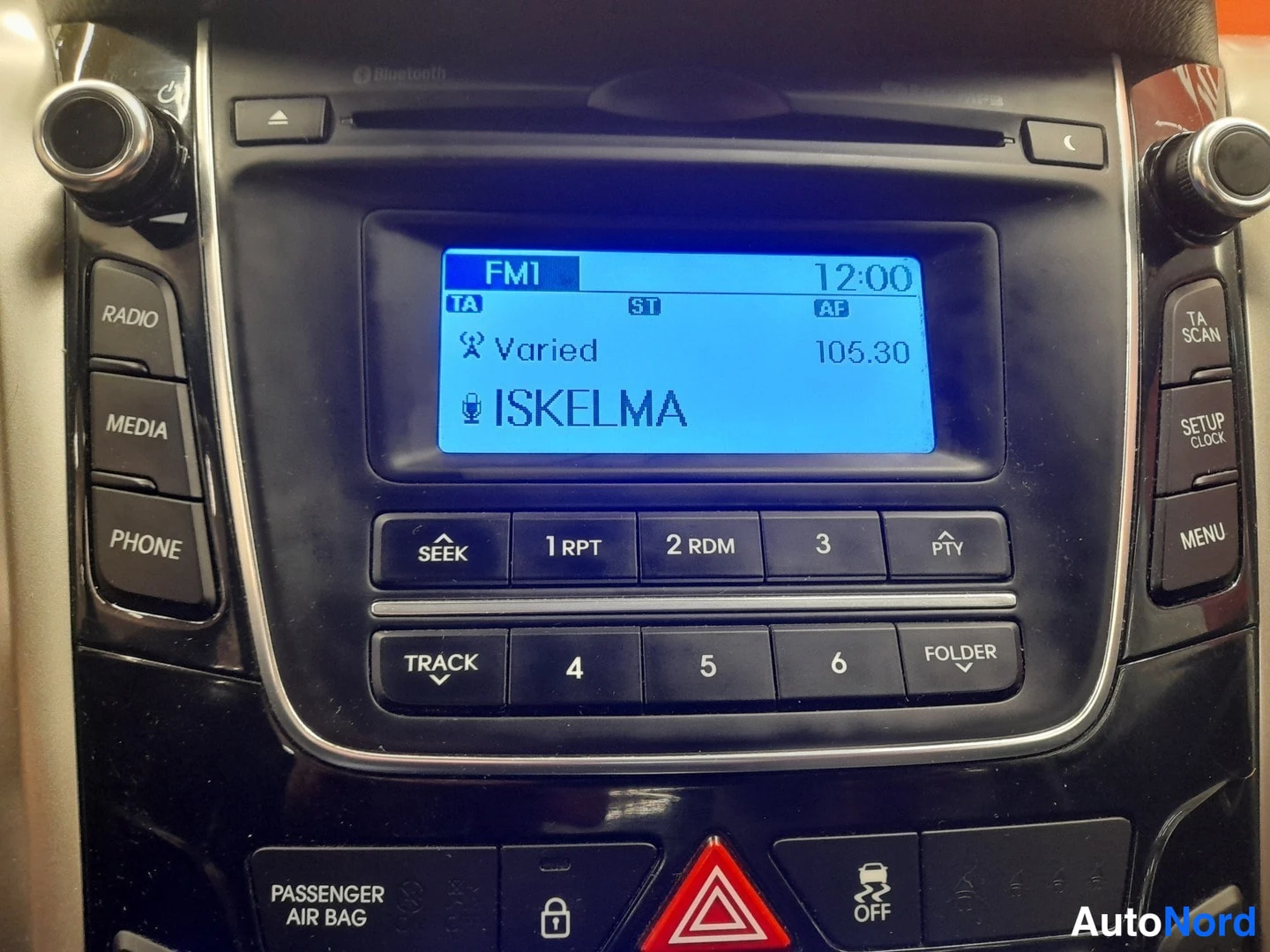 Edestä: Radiopanelin tarkempi kuva: näytössä FM1 105,30 Iskelma, kello 12:00, ja näppäimet RADIO, MEDIA, PHONE, SEEK, TRACK ja FOLDER.