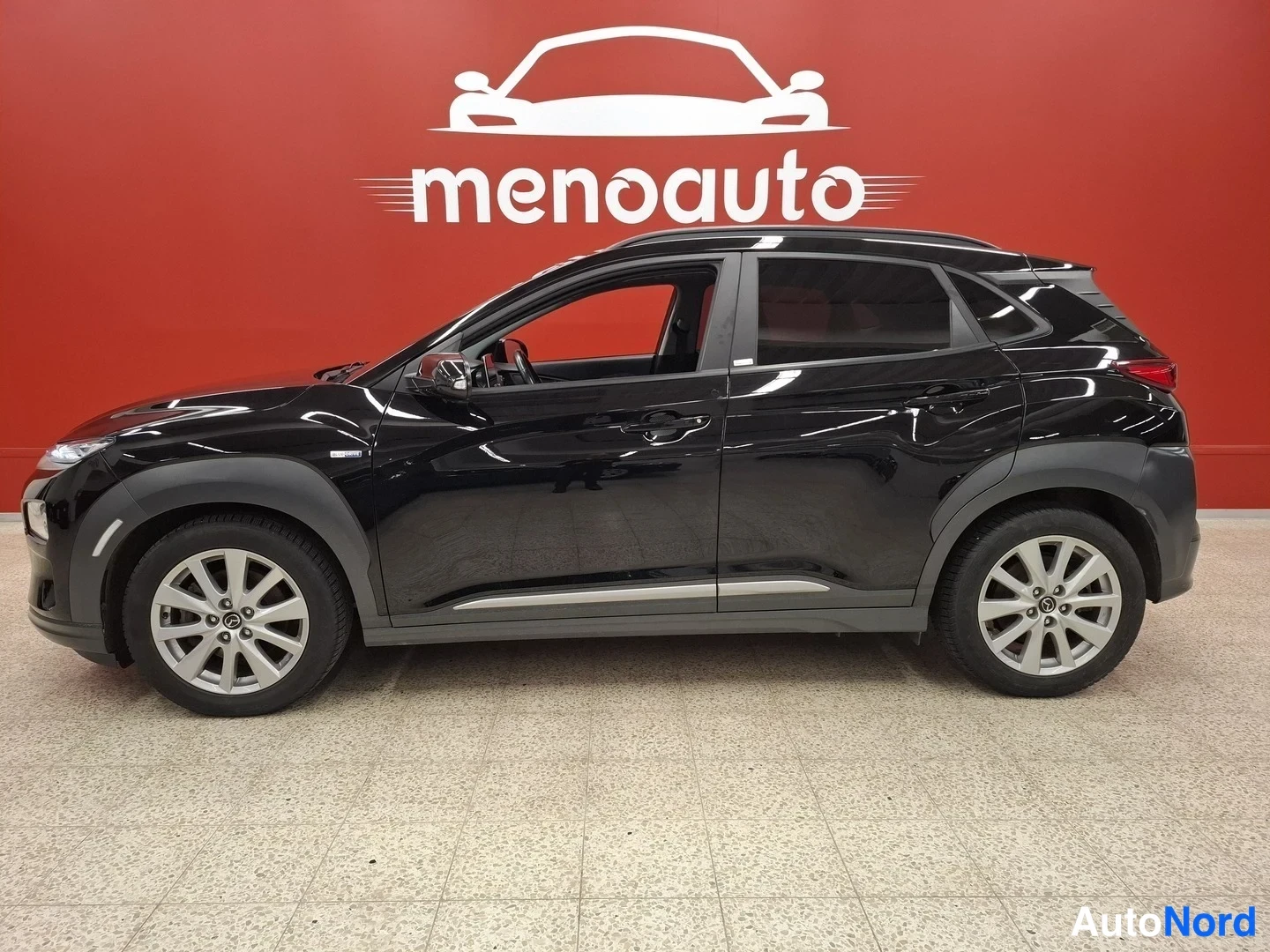 Sivulta: Musta Hyundai Kona-electric-hatchback sivulta kuvattuna sisätilassa, punainen menoauto-taustaseinä.