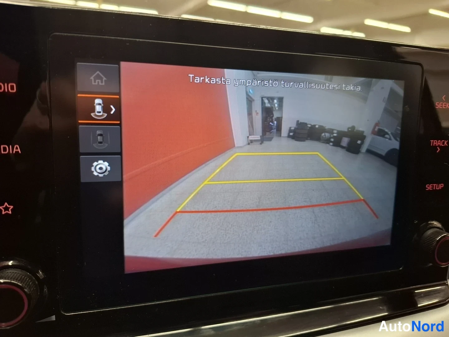 Yksityiskohta: Infotainment-näyttö, jolla näkyy peruutuskameran kuva taksipylväineen ja ohjausviivoineen sekä suomenkielinen varoitusteksti.