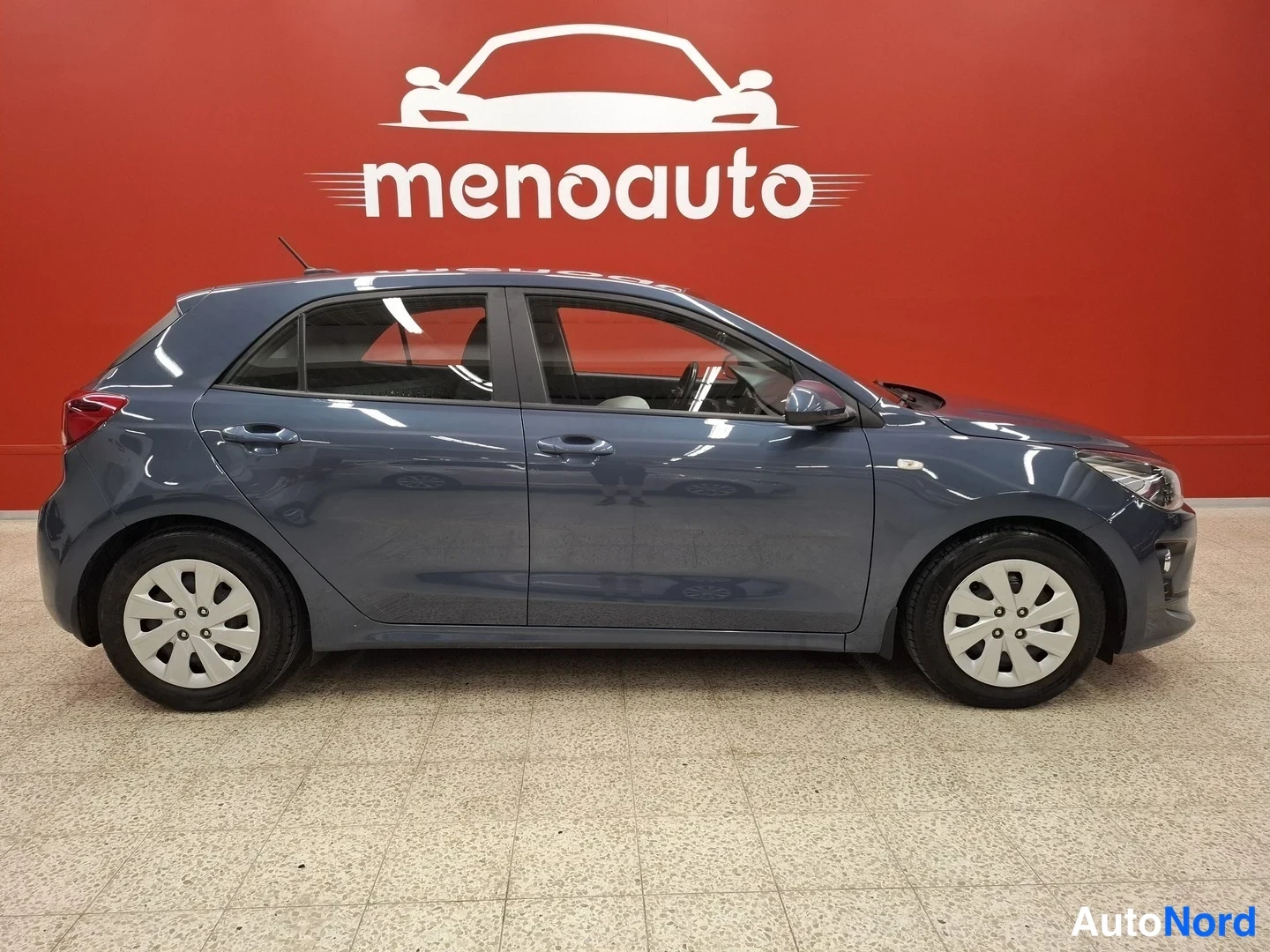 Sivulta: Sinisenharmaan Kia Rio -hatchbackin sivuprofiili vasemmalta kuvattuna autohallissa punaisella taustalla.