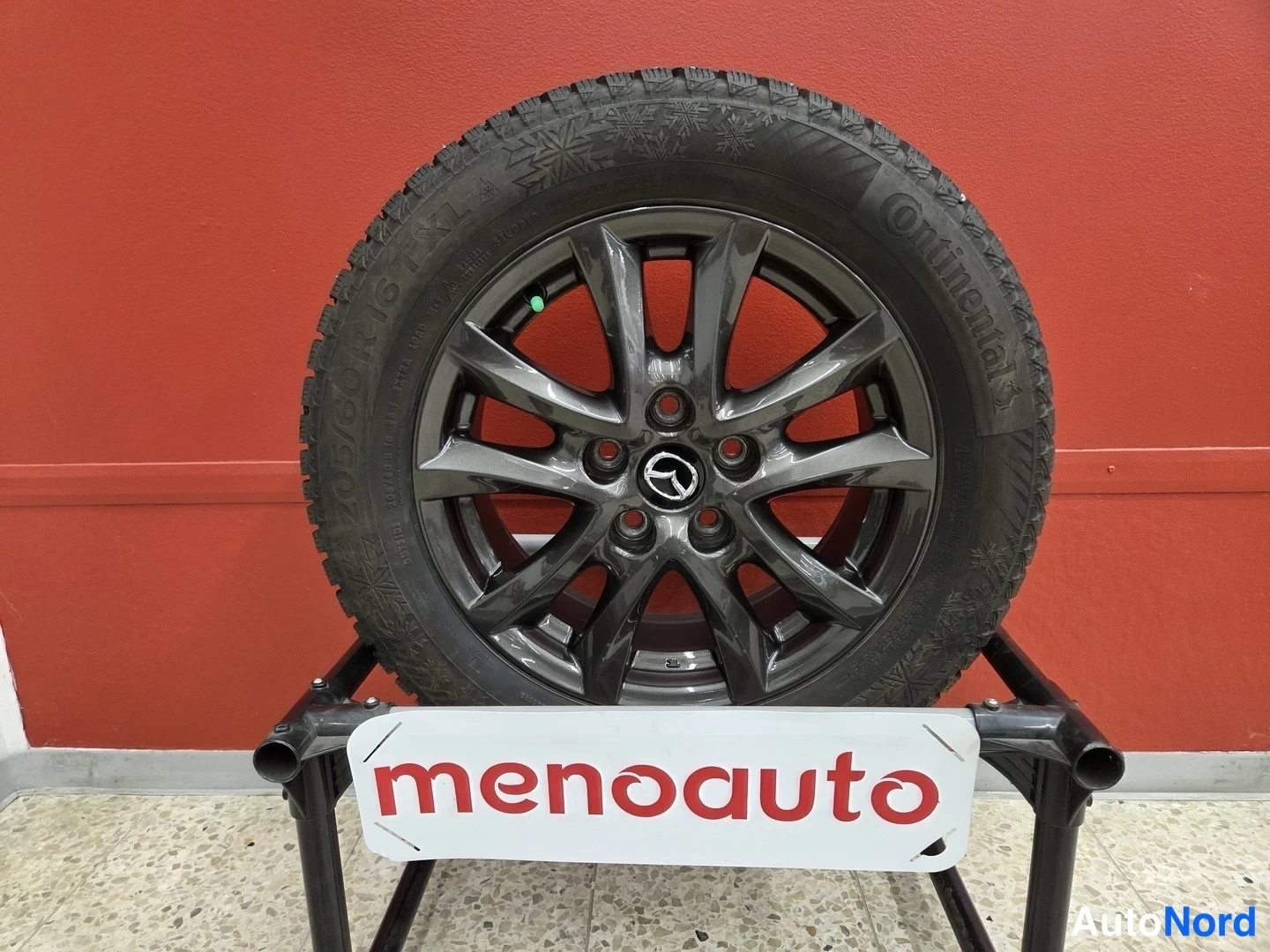 Yksityiskohta: Continental-talvirengas (205/60 R16) tummalla Mazda-vanteella jalustalla punaisella seinällä, jalustassa menoauto-tunnus.