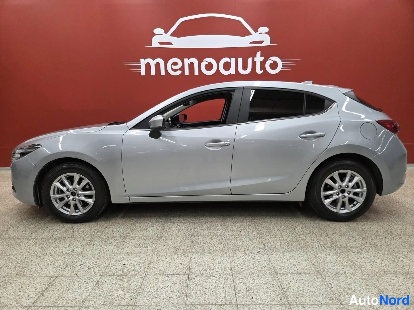 Sivulta: Hopeanvärainen Mazda 3 -viistoperä sivulta kuvattuna sisätiloissa, vasen kylki näkyvissä.