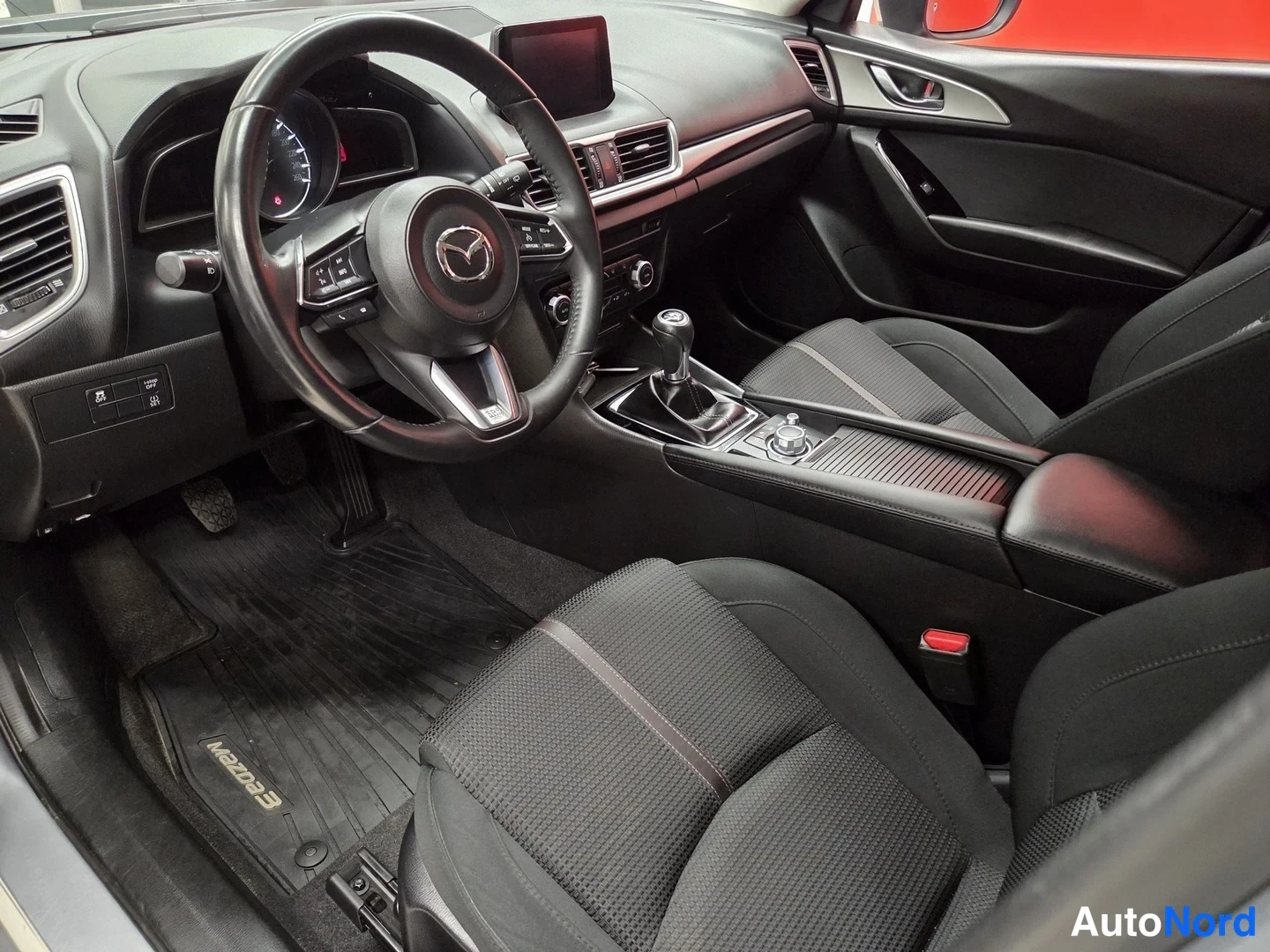 Muu: Mazda 3:n ohjaamo kuljettajan puolelta: mustat penkit, manuaalivaihtajan keppi ja Mazda-merkitty lattiamatto.