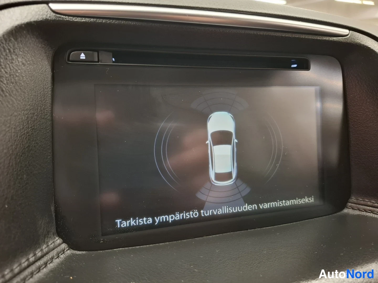 Muu: Keskikonsolin näyttö, jossa kamera- tai tutkakuva autosta ja teksti 'Tarkista ympäristö turvallisuuden varmistamiseksi'.